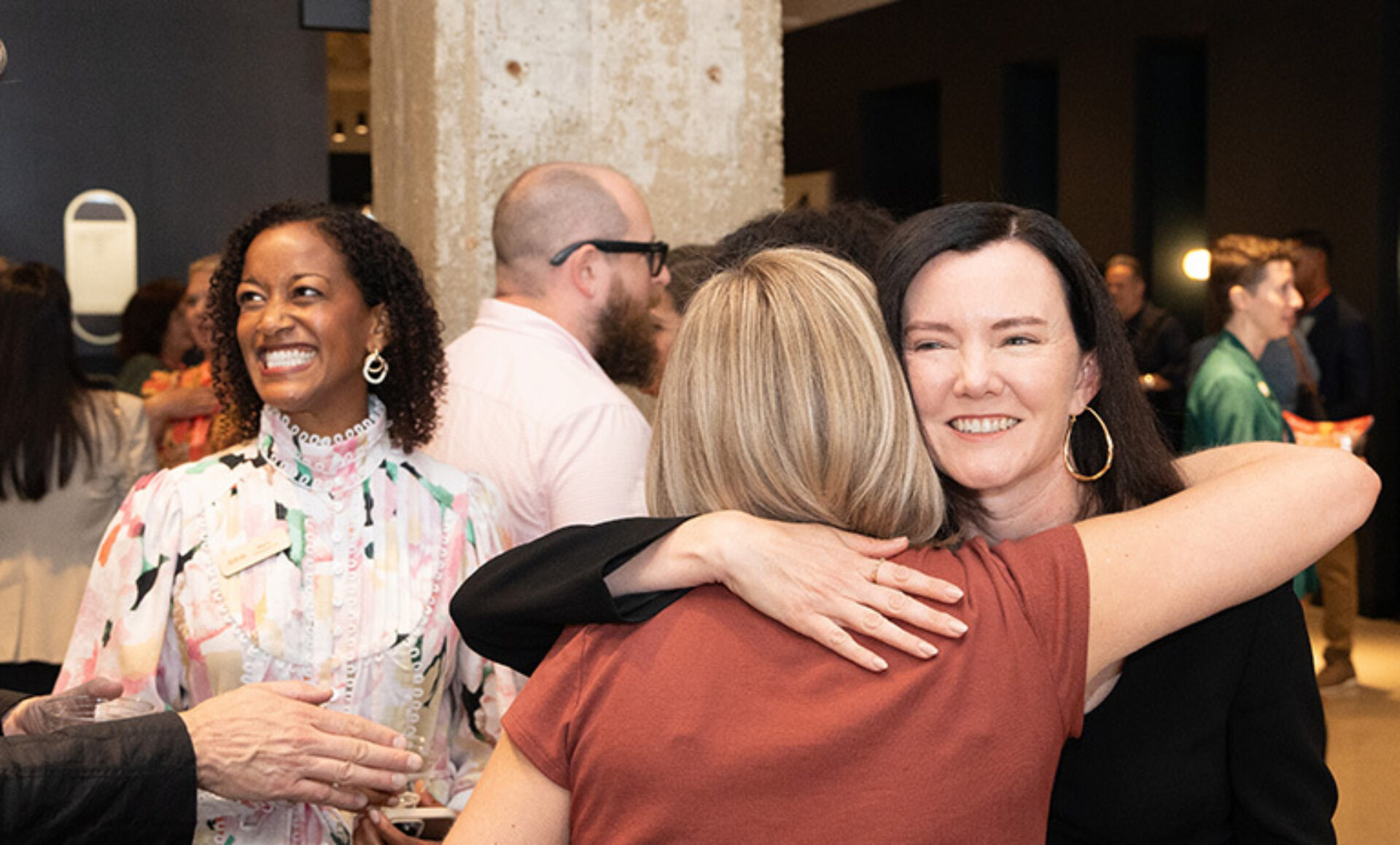 Woman with dark hair and hoop earrings embraces a blonde woman while a smiling woman in a pastel floral blouse watches at a busy reception.