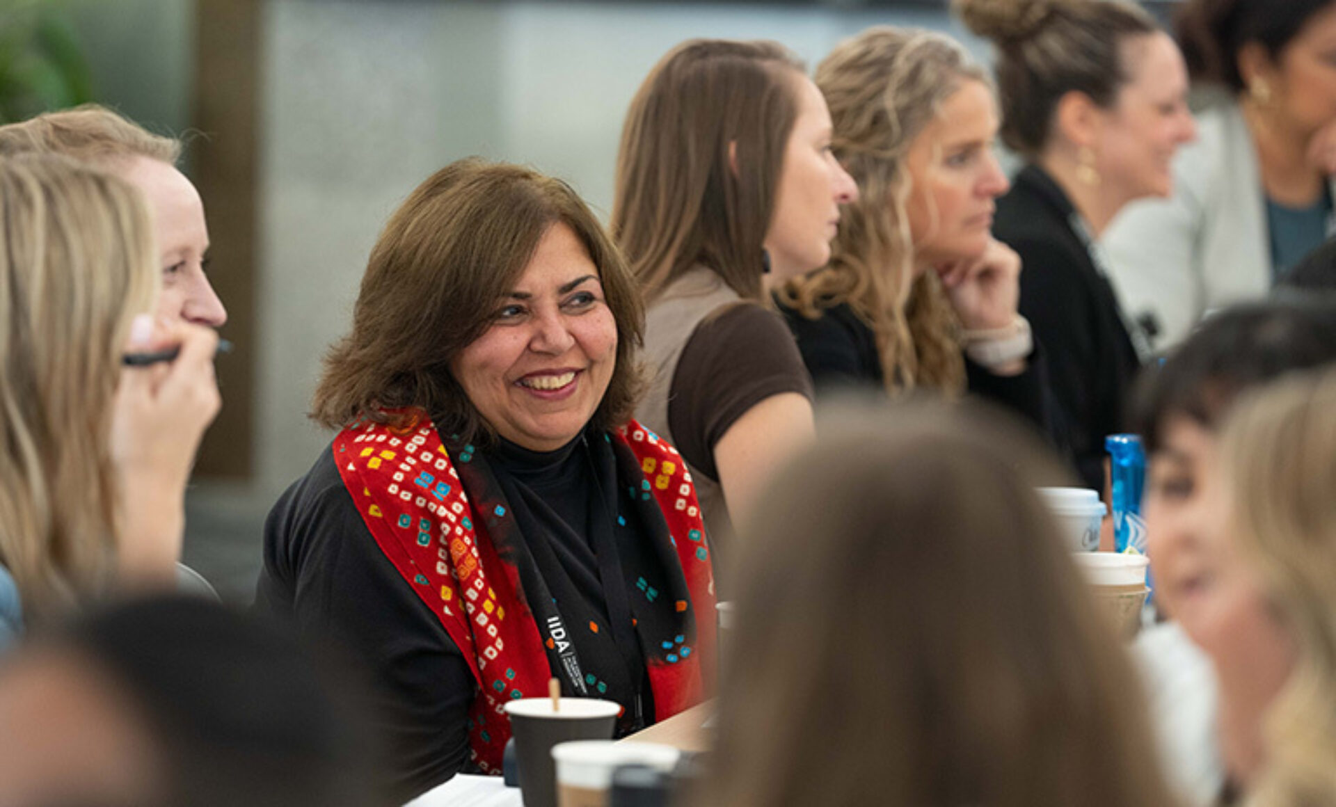 A woman with a warm smile wears a colorful patterned scarf, engaged in a lively conversation with a group of people at a table.