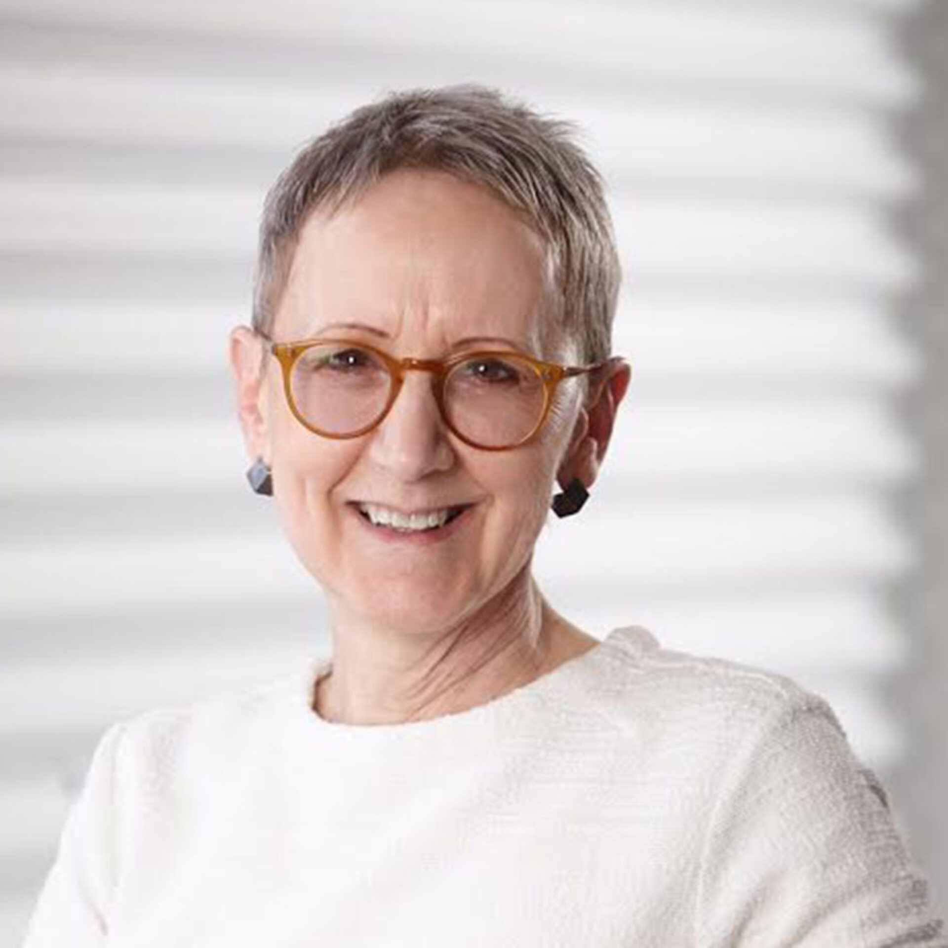 Smiling older woman with short gray hair and round amber glasses, wearing black geometric earrings and a white textured top against blinds.