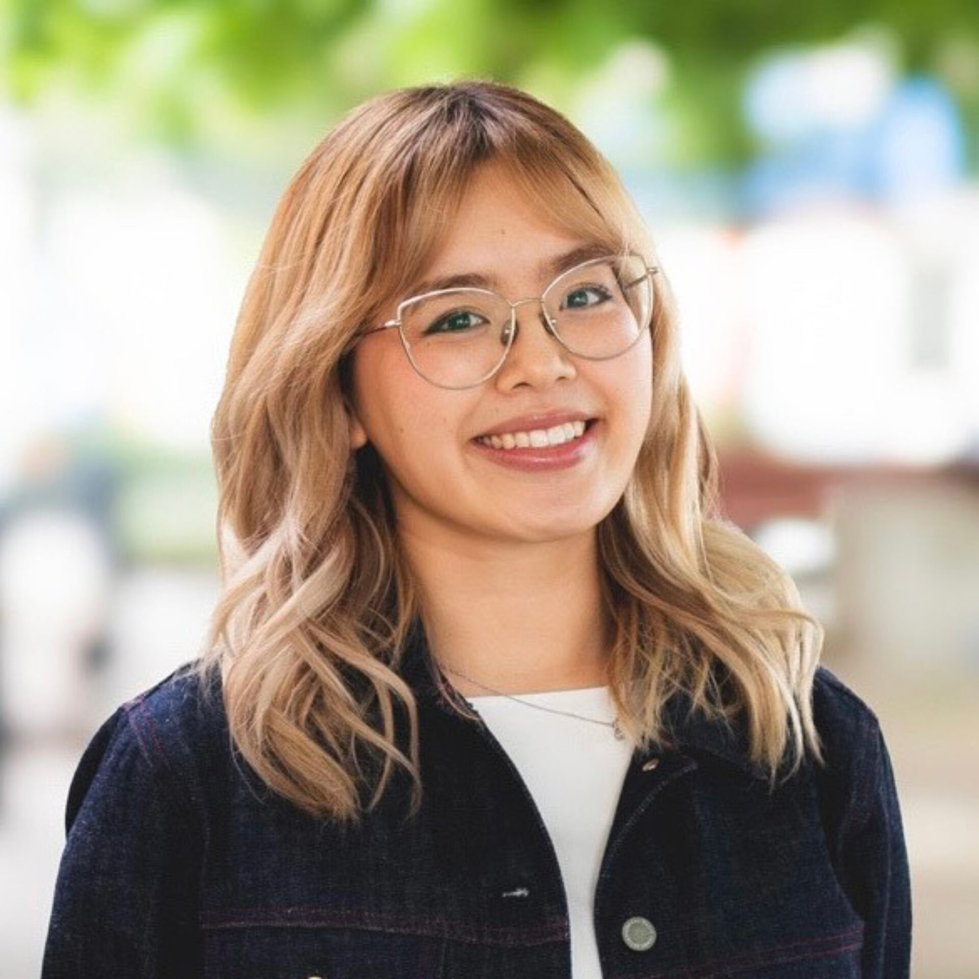 Young woman with shoulder-length blonde hair and glasses smiling, wearing a dark jacket over a white top with blurred green background.