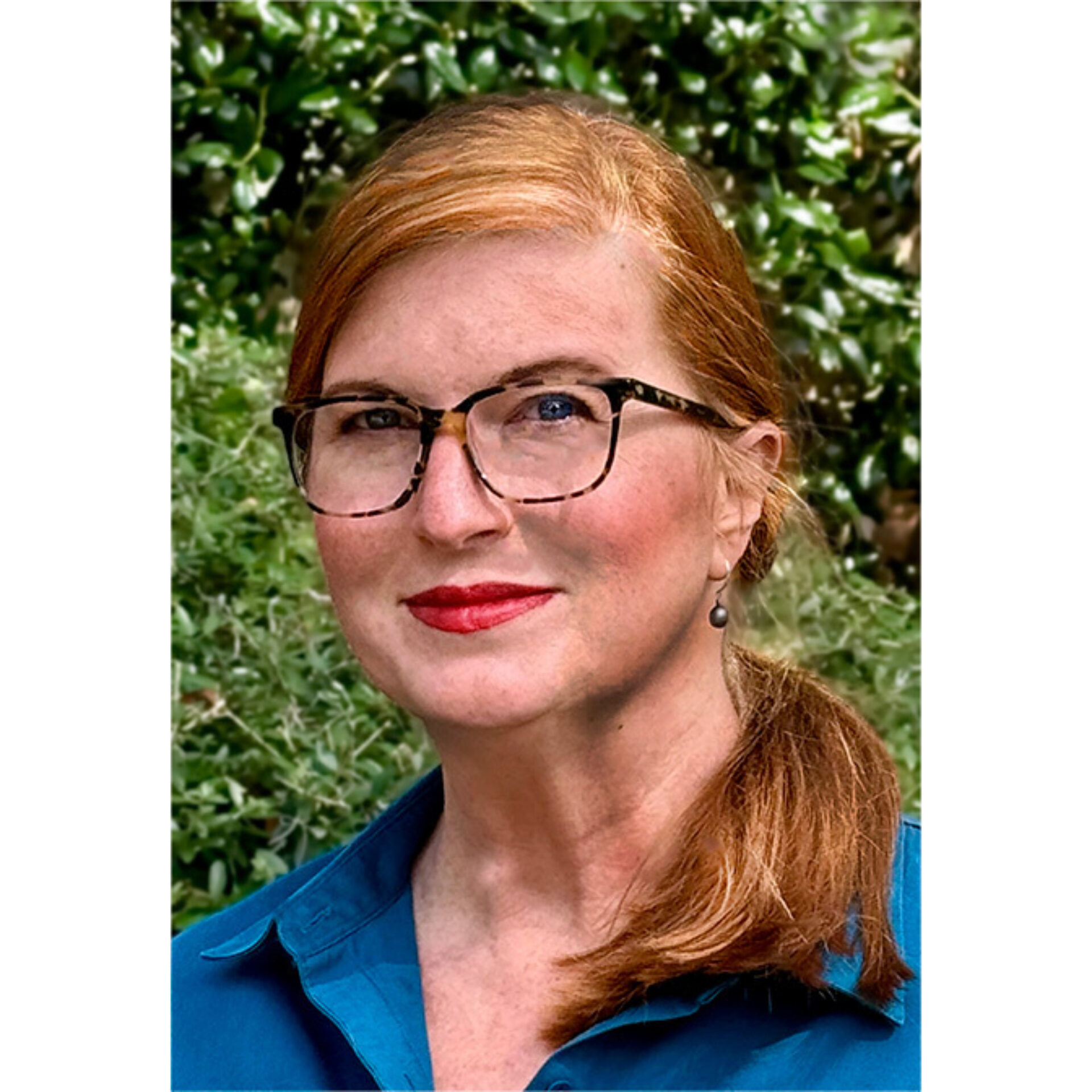 Red-haired woman wearing tortoise glasses and red lipstick, blue collared shirt, drop earring, hair in a low ponytail, green foliage.