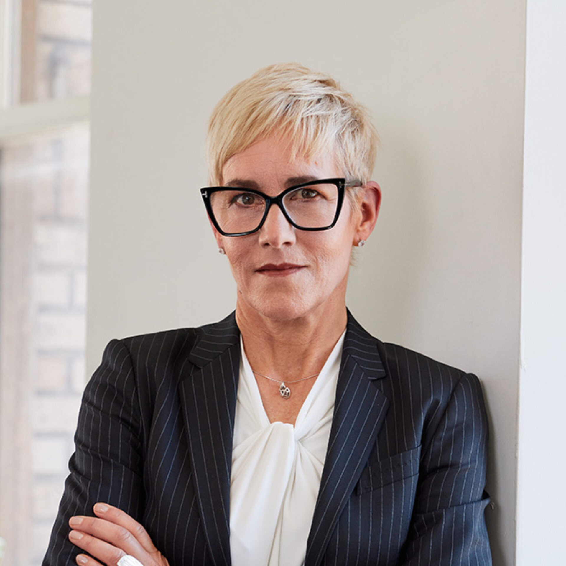 Short platinum hair, black angular glasses, pinstripe navy blazer over a white tied blouse, arms crossed while leaning against a pale wall.