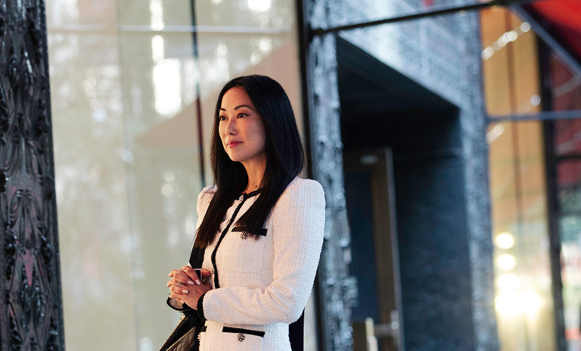 A woman with long black hair stands thoughtfully in a stylish, modern setting, wearing a chic white tweed outfit and holding a handbag.