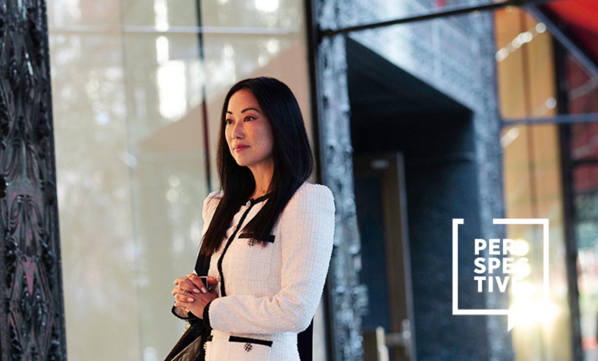 A woman with long black hair, wearing a stylish white suit, stands thoughtfully by large glass doors in a modern setting.