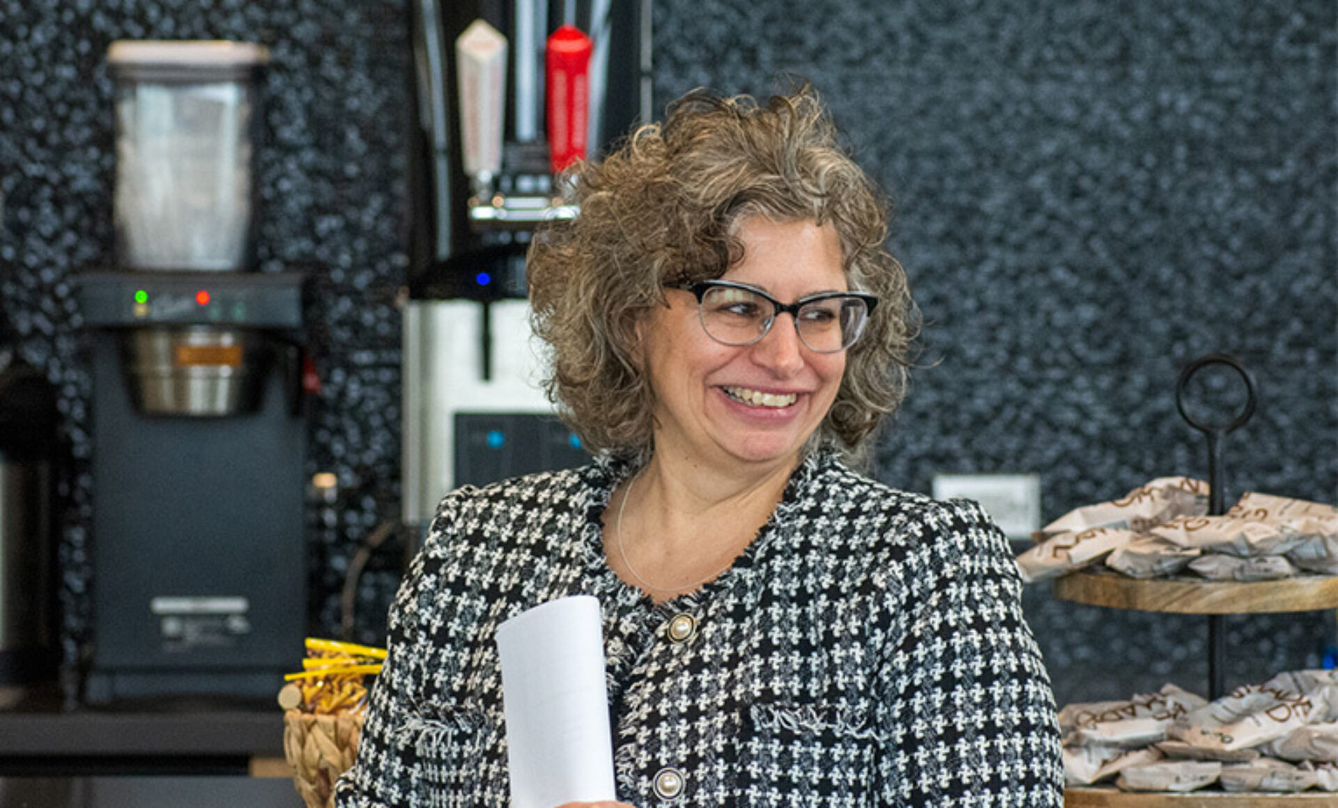 Smiling woman with curly gray hair and glasses in a black-and-white houndstooth jacket holds a rolled paper in a café setting.