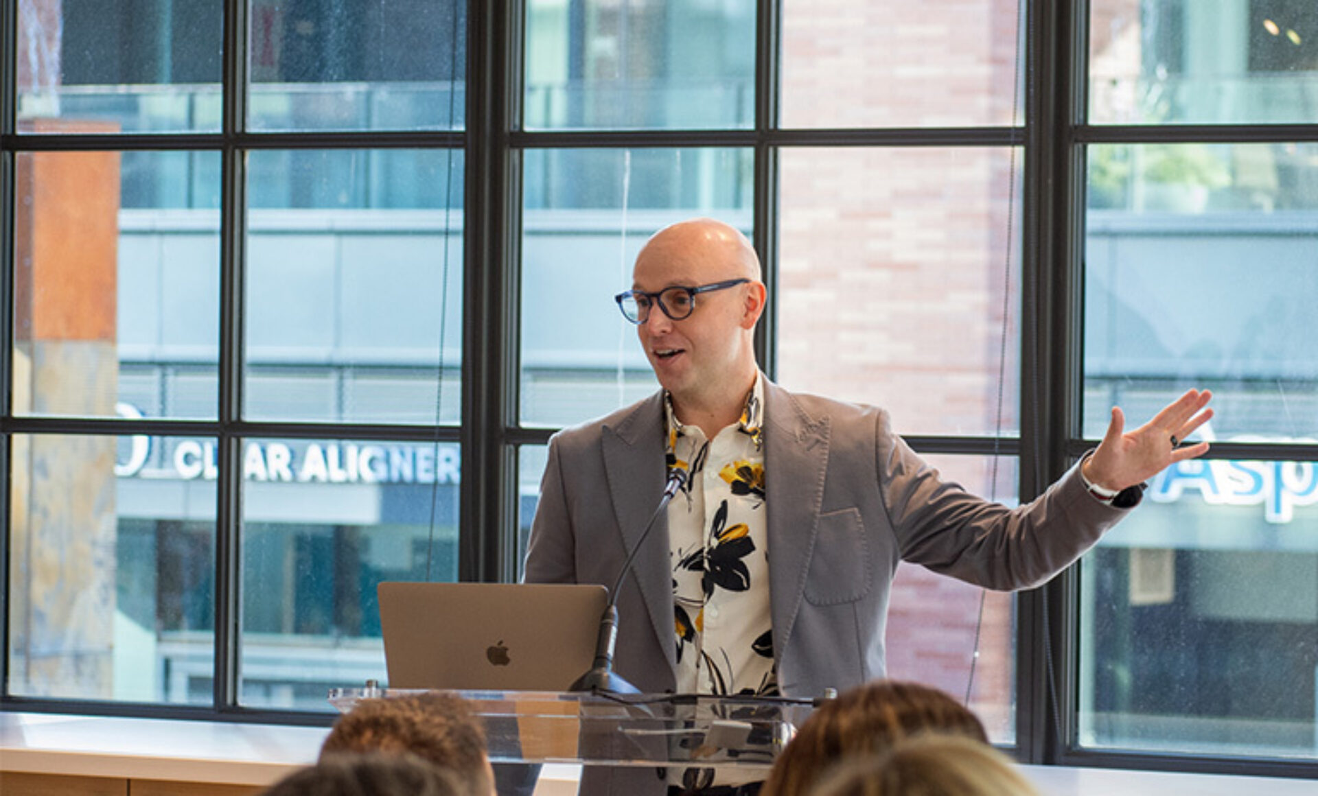 A speaker in a stylish floral shirt gestures enthusiastically while presenting in front of a laptop, engaging an attentive audience.