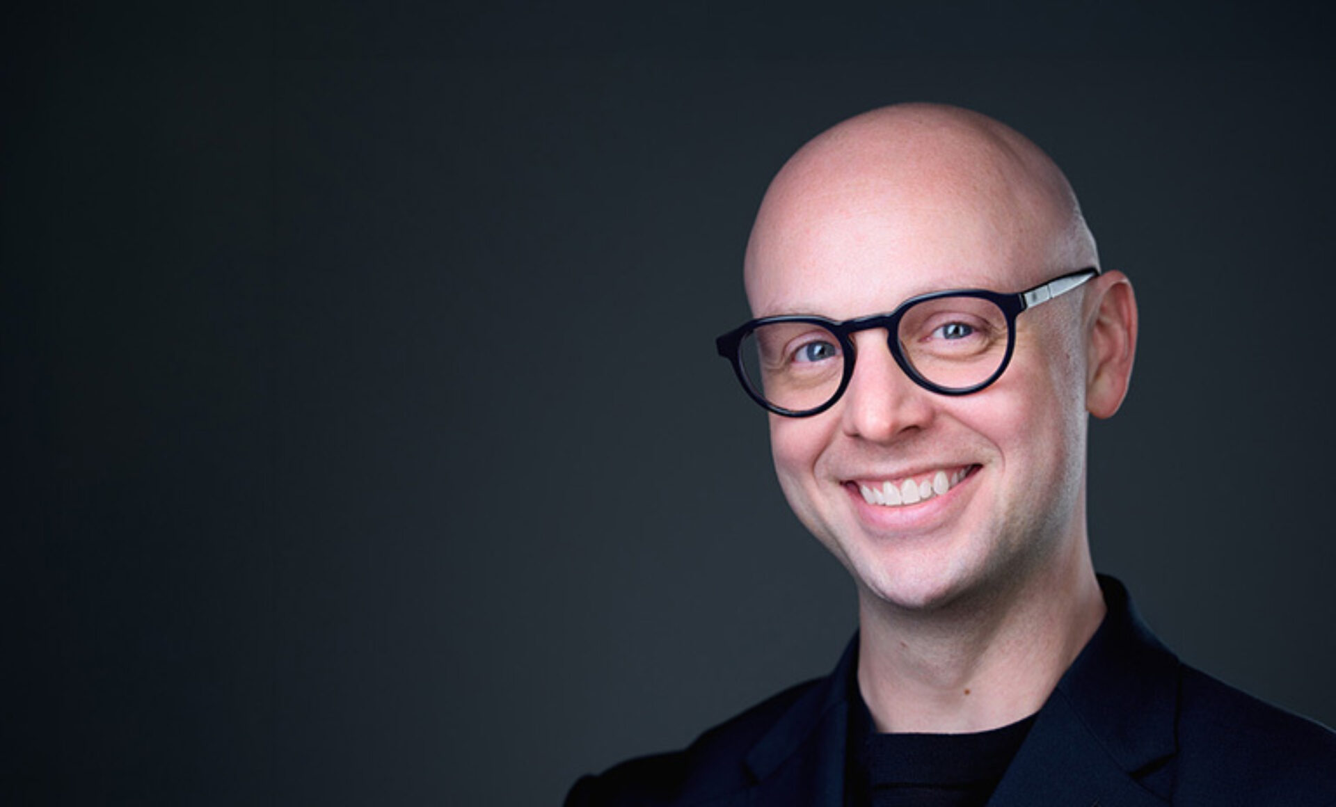 Smiling bald man in round black glasses and a dark blazer against a soft dark gray background, head tilted slightly to the right.