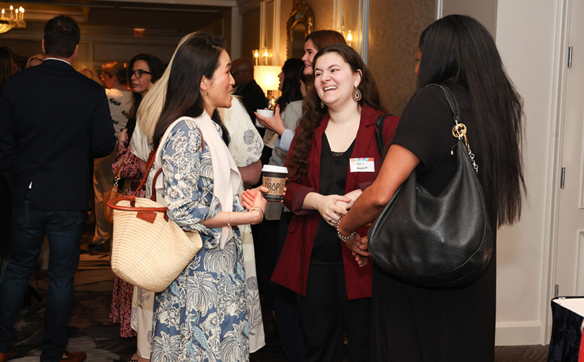 Three women chat at a networking event: blue print dress with straw tote, burgundy blazer holding coffee, and black dress with leather bag.