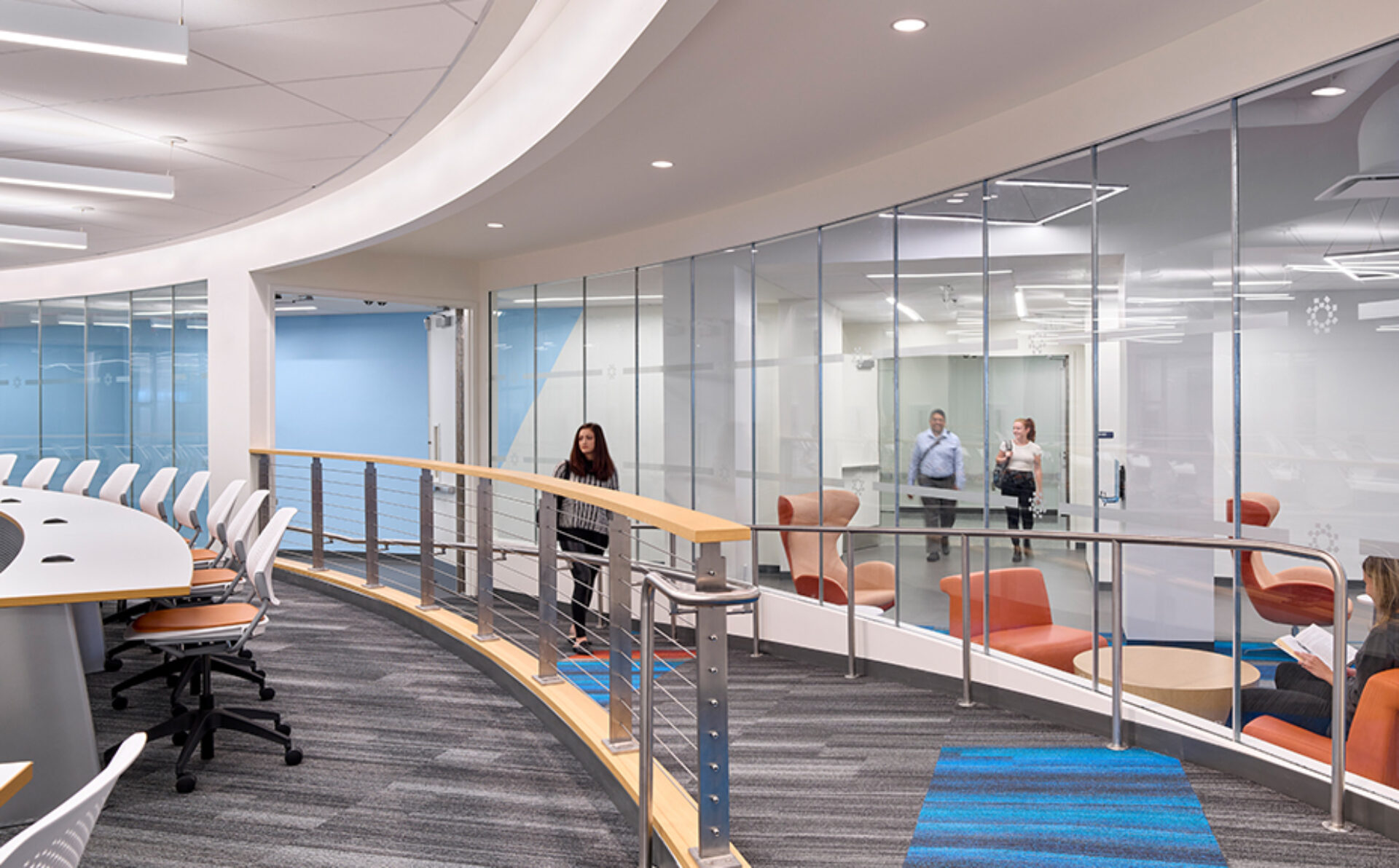 Curved modern office corridor with glass-walled meeting rooms, orange lounge chairs, a woman walking along the rail and people inside rooms.