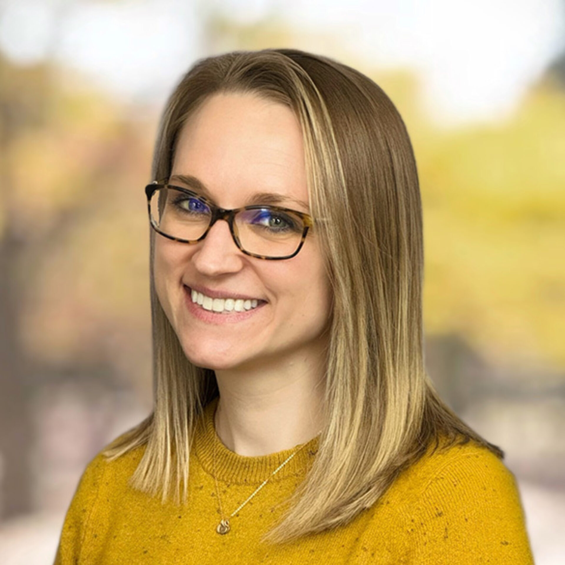 Smiling woman with shoulder-length blonde hair, tortoiseshell glasses and mustard sweater wearing a pendant, blurred autumn background.