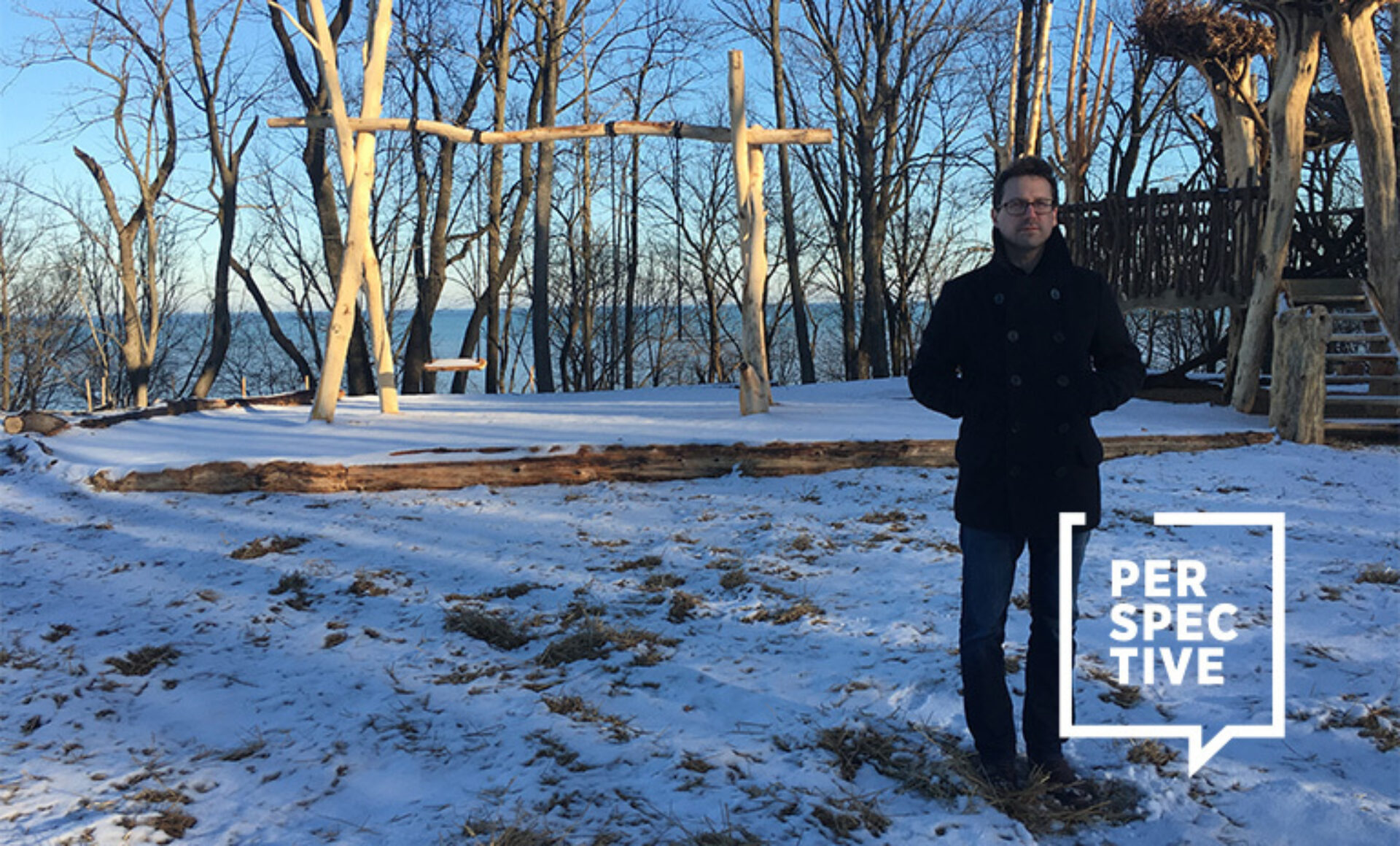A man stands in a snowy landscape surrounded by trees, with wooden structures in the background and a distant view of water.