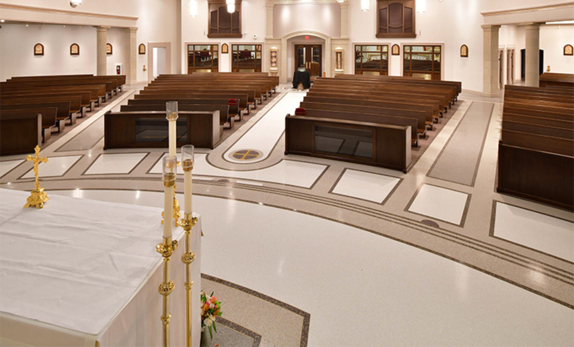 Bright Catholic church interior with central aisle flanked by wooden pews, altar foreground with tall white candles, gold crucifix, flowers.