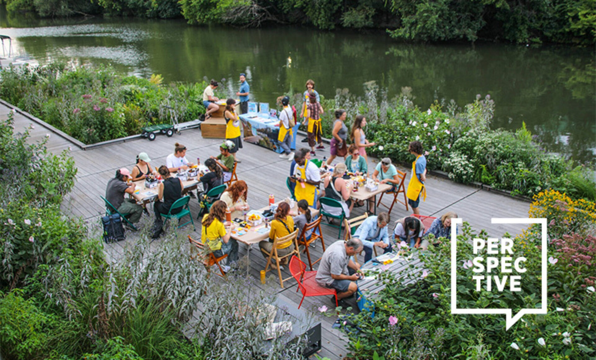 A vibrant gathering along a river, with people enjoying food, surrounded by lush greenery and colorful flowers on a wooden deck.