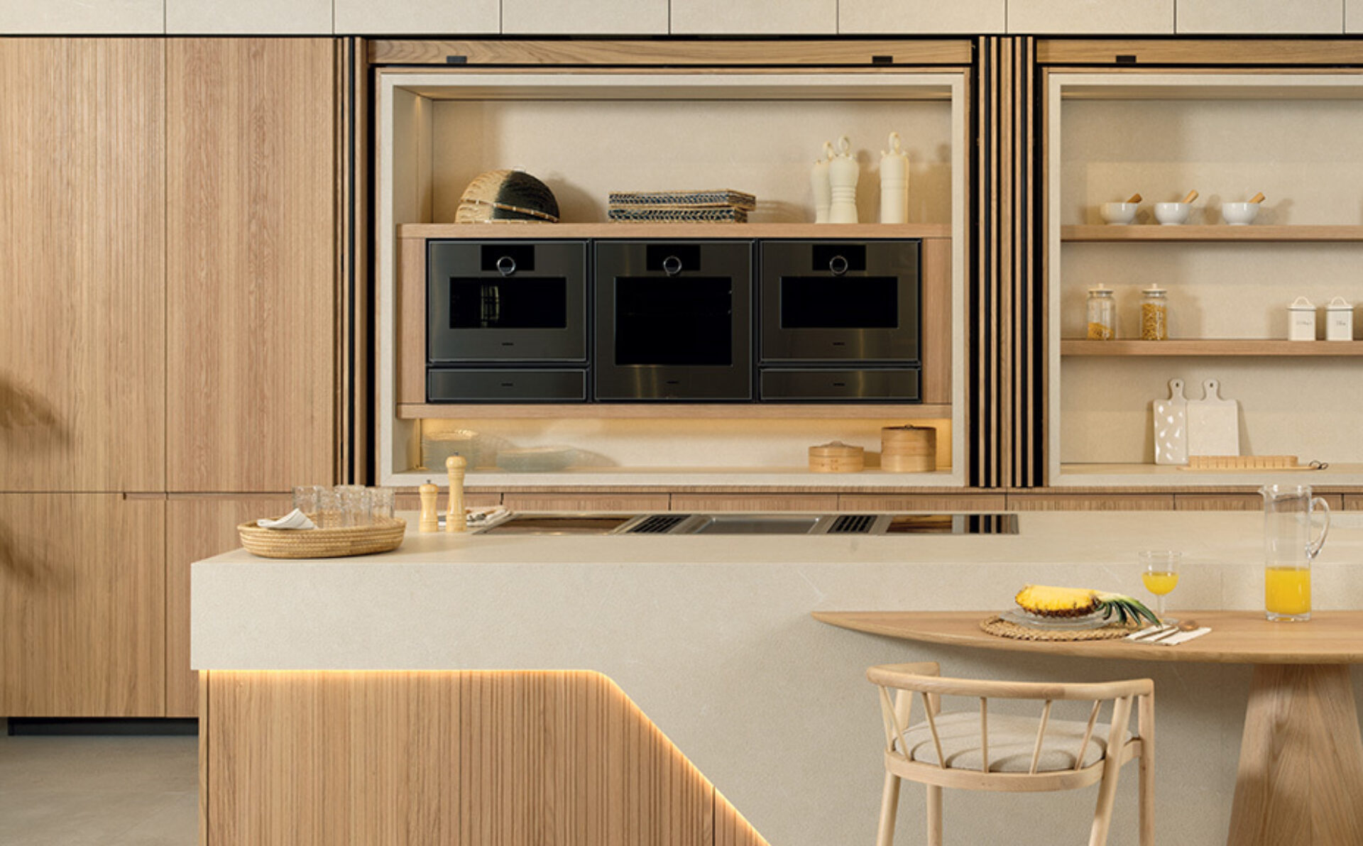 Minimalist warm wood kitchen with three black built-in ovens, open shelves, a stone island with cooktop, tray, pineapple and juice.