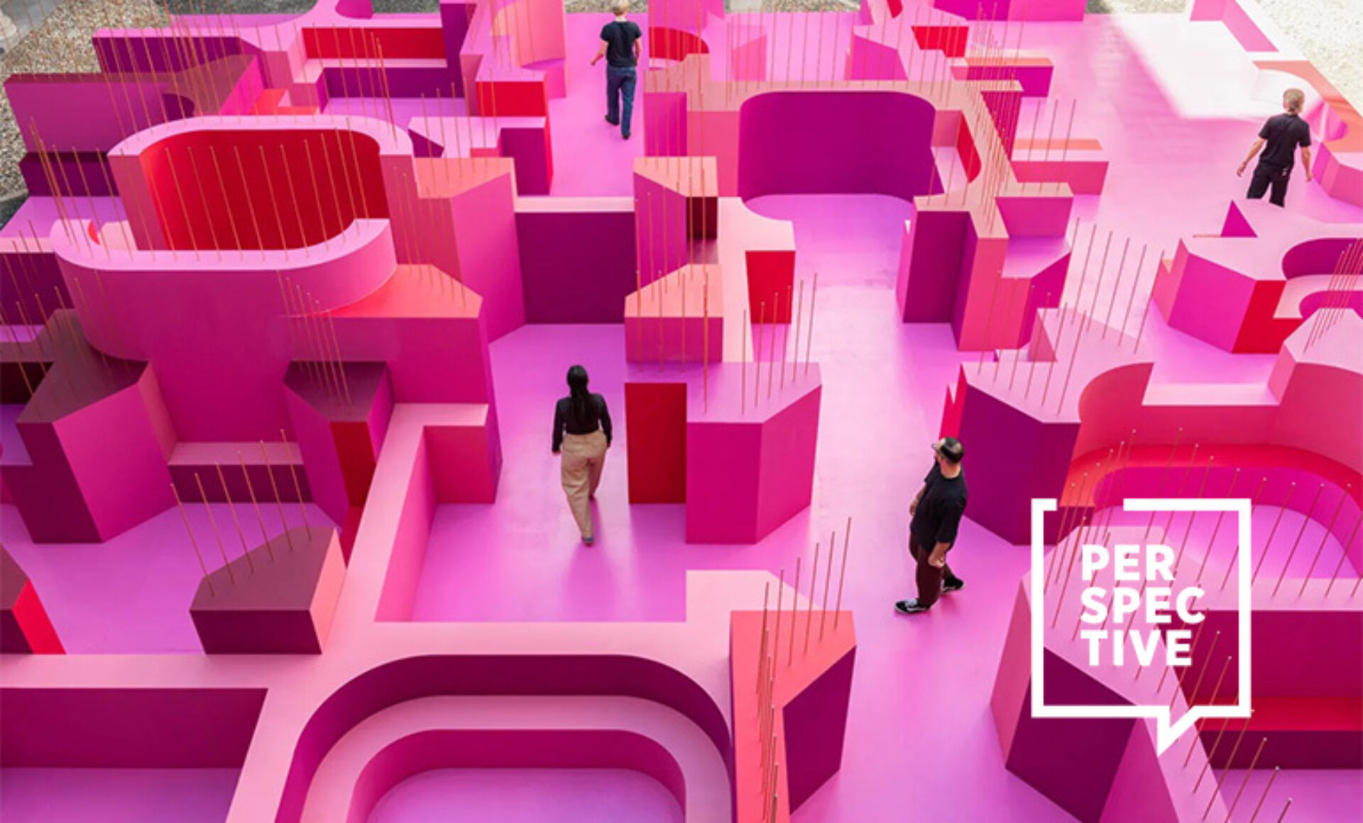 Aerial view of a bright magenta modular maze installation with curved walls and tall thin rods, five people walking through its levels.