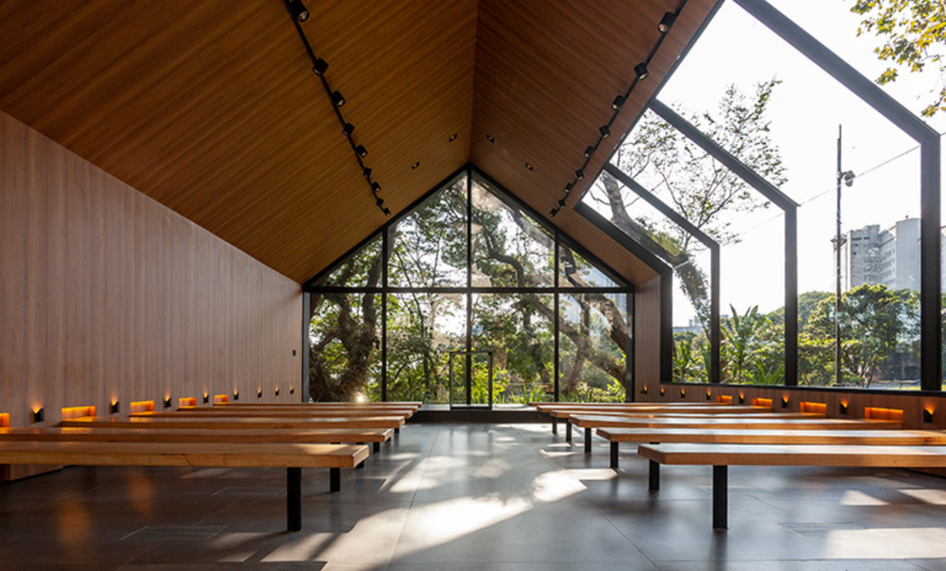 Minimal chapel interior with A-frame wood ceiling, wooden benches, and floor-to-ceiling glass revealing trees and warm sunlight.
