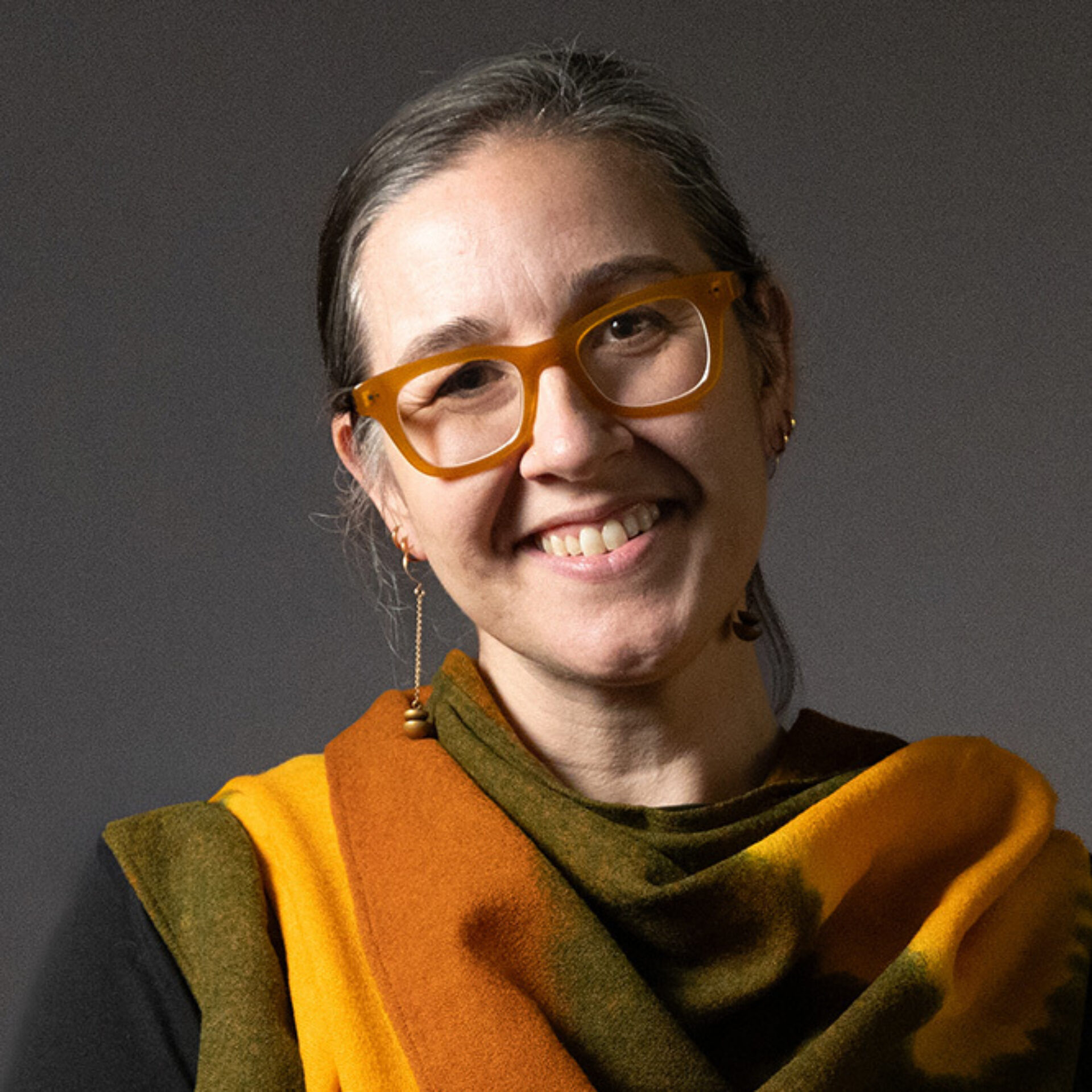 Smiling woman with gray hair pulled back, mustard-yellow glasses and dangling earring, wearing orange-green shawl over a dark top.