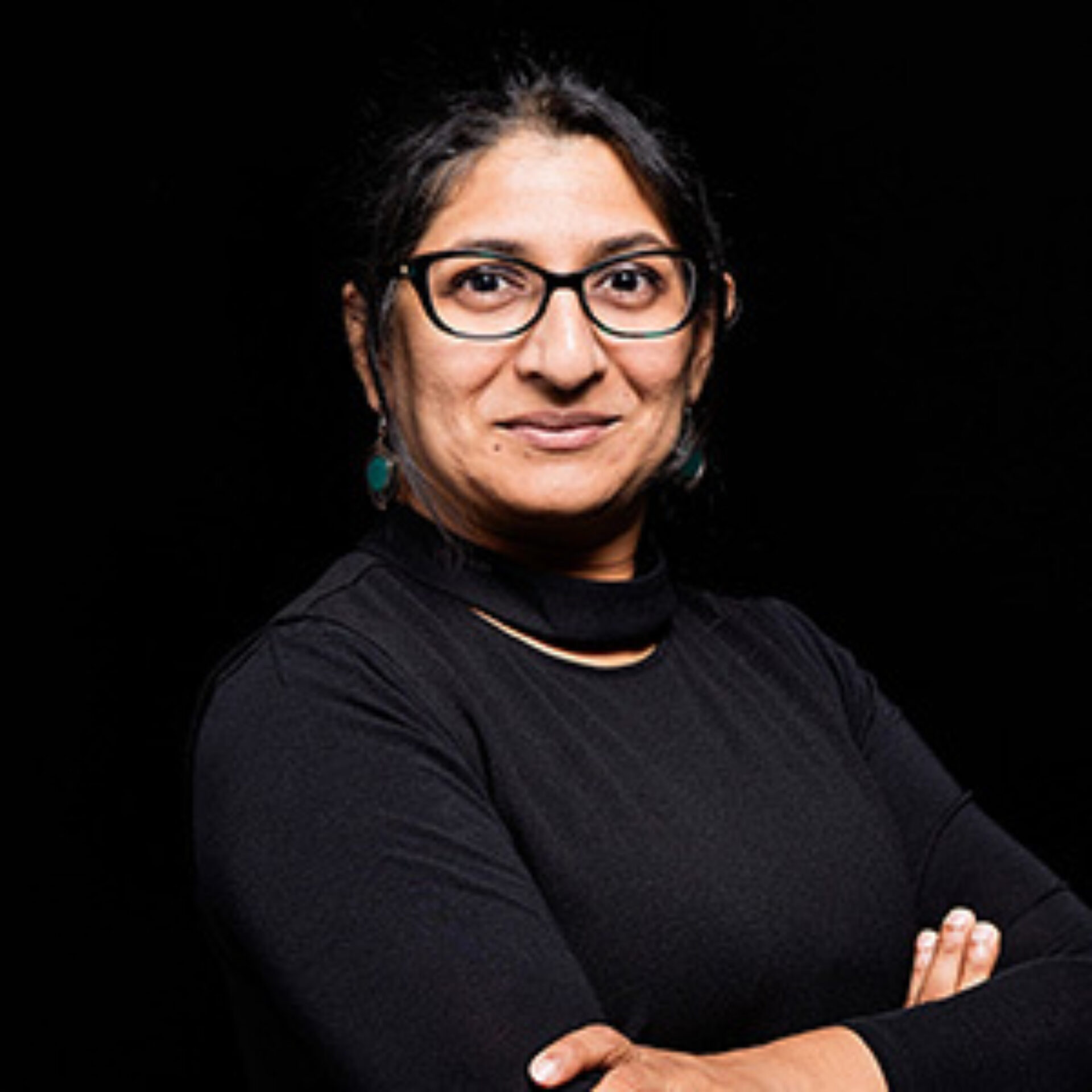 Shoulder-length portrait of a woman with dark hair in a bun, black glasses and turtleneck, turquoise earrings, arms crossed, subtle smile.