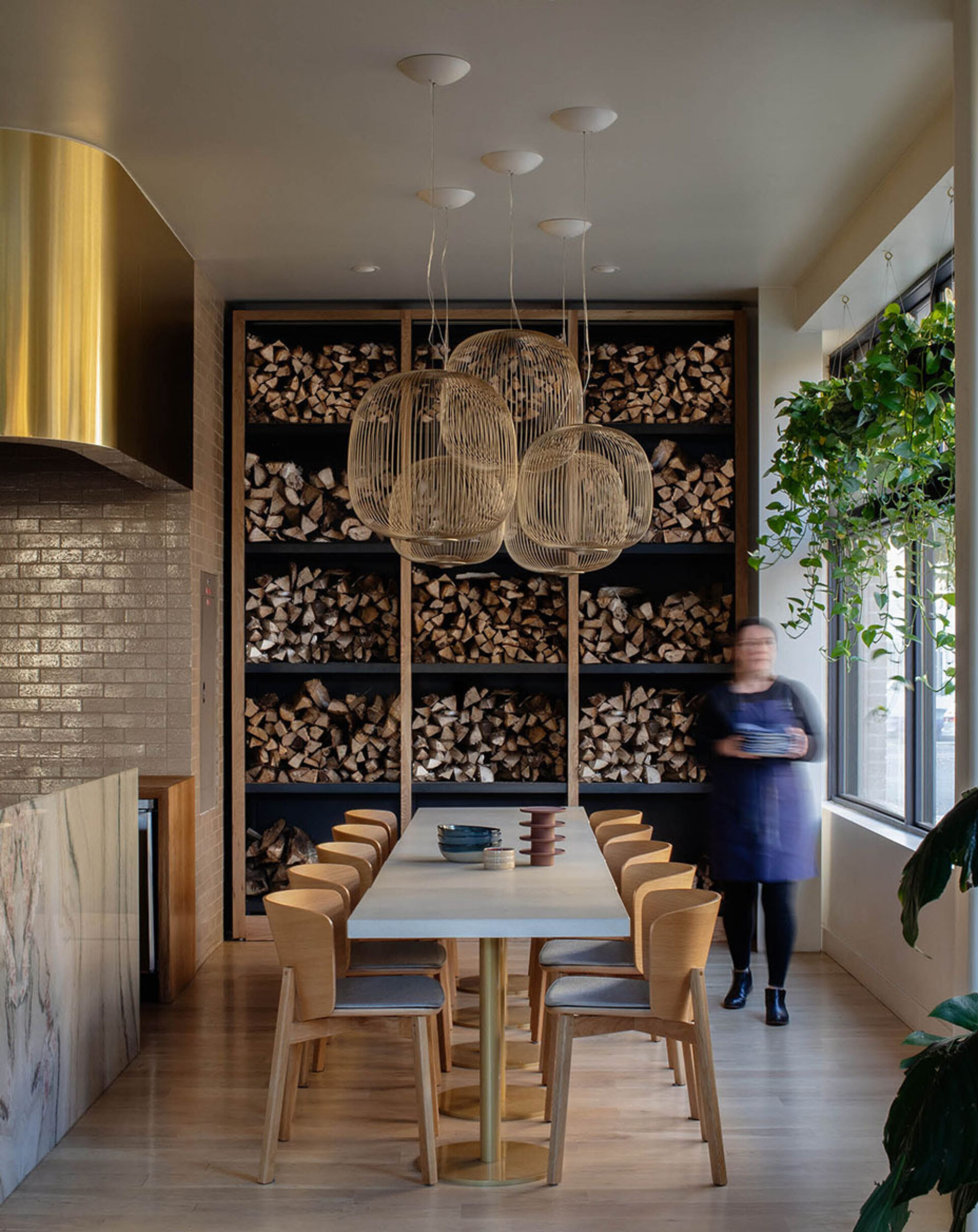 A modern dining area features a long table surrounded by wooden chairs, with stacked firewood in the background and pendant lighting overhead.