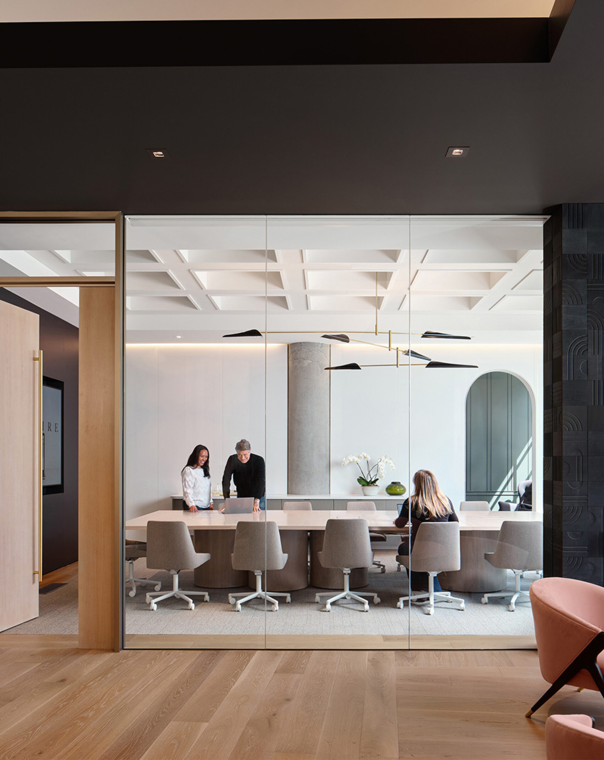 A modern conference room featuring a large table, stylish chairs, and three people discussing around it, with natural light illuminating the space.