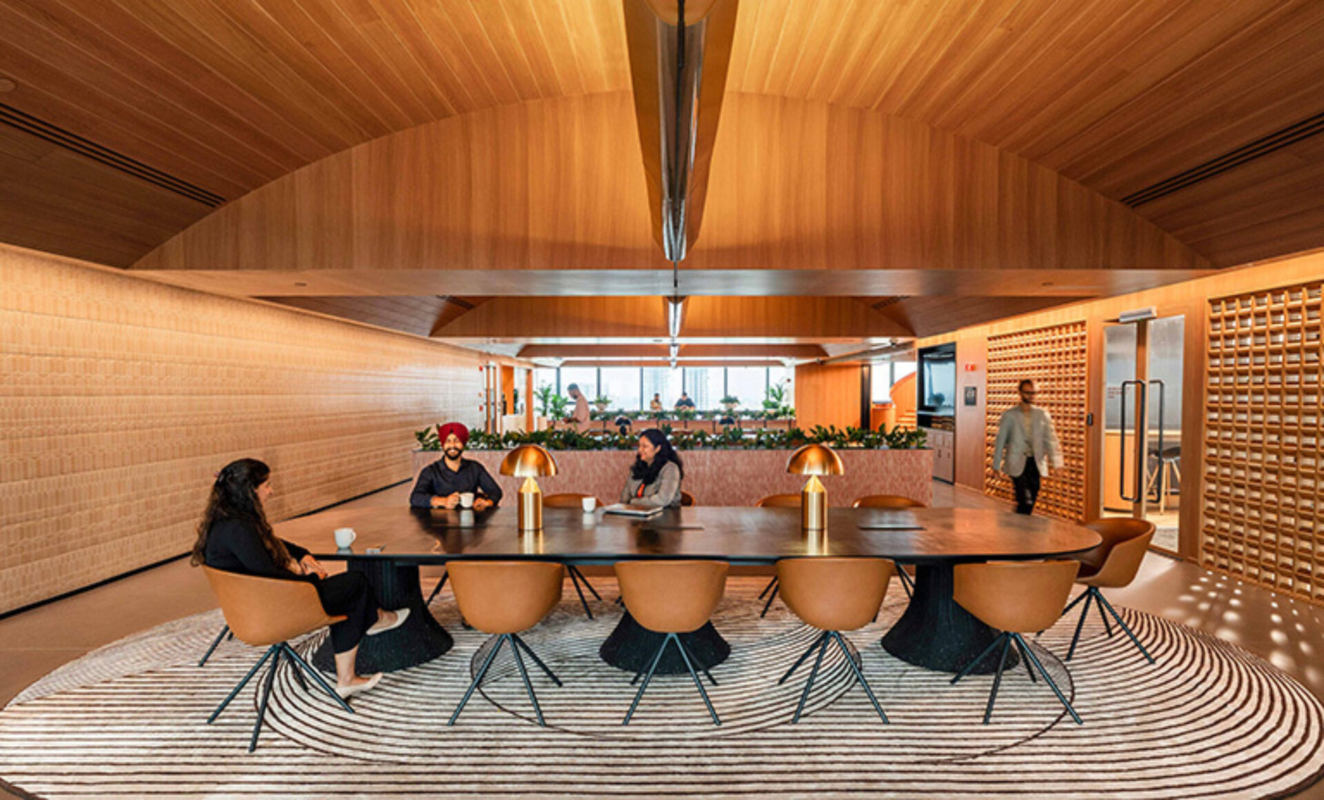 A modern conference room features a large table surrounded by stylish chairs, with individuals engaged in discussion and plants in the background.