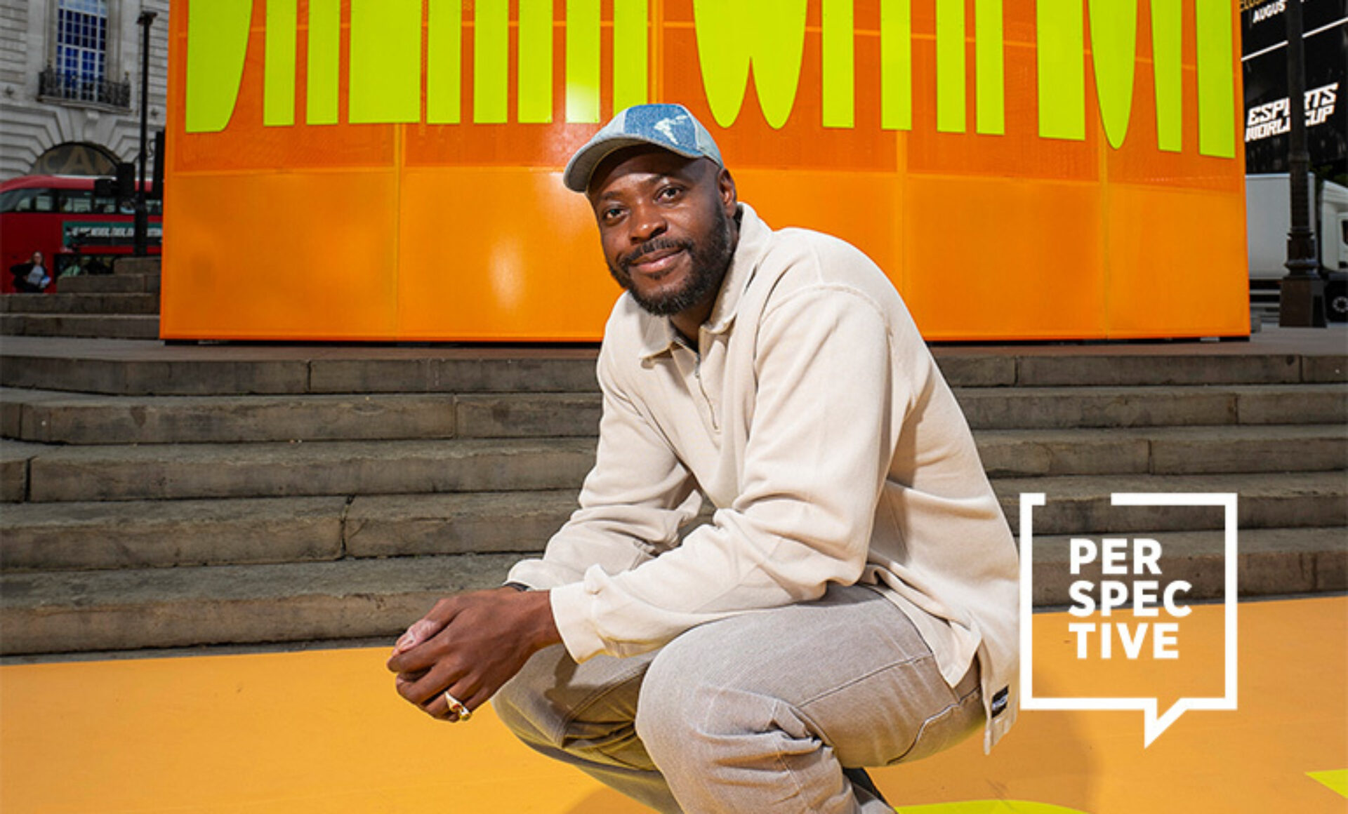 Man crouches on yellow floor before an orange panel with large green letters, wearing beige sweatshirt and cap, 'PERSPECTIVE' logo lower right.