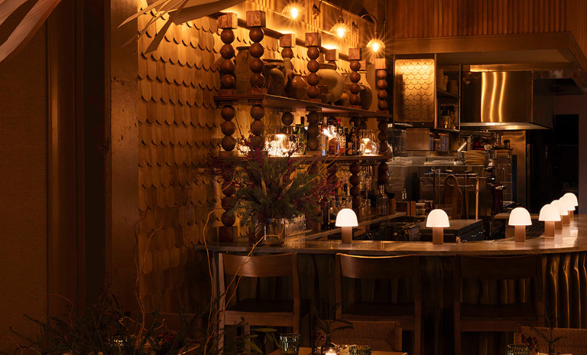 Warm, dimly lit bar with a curved wooden counter, row of small mushroom lamps, high stools, ornate shelving with bottles and beads.