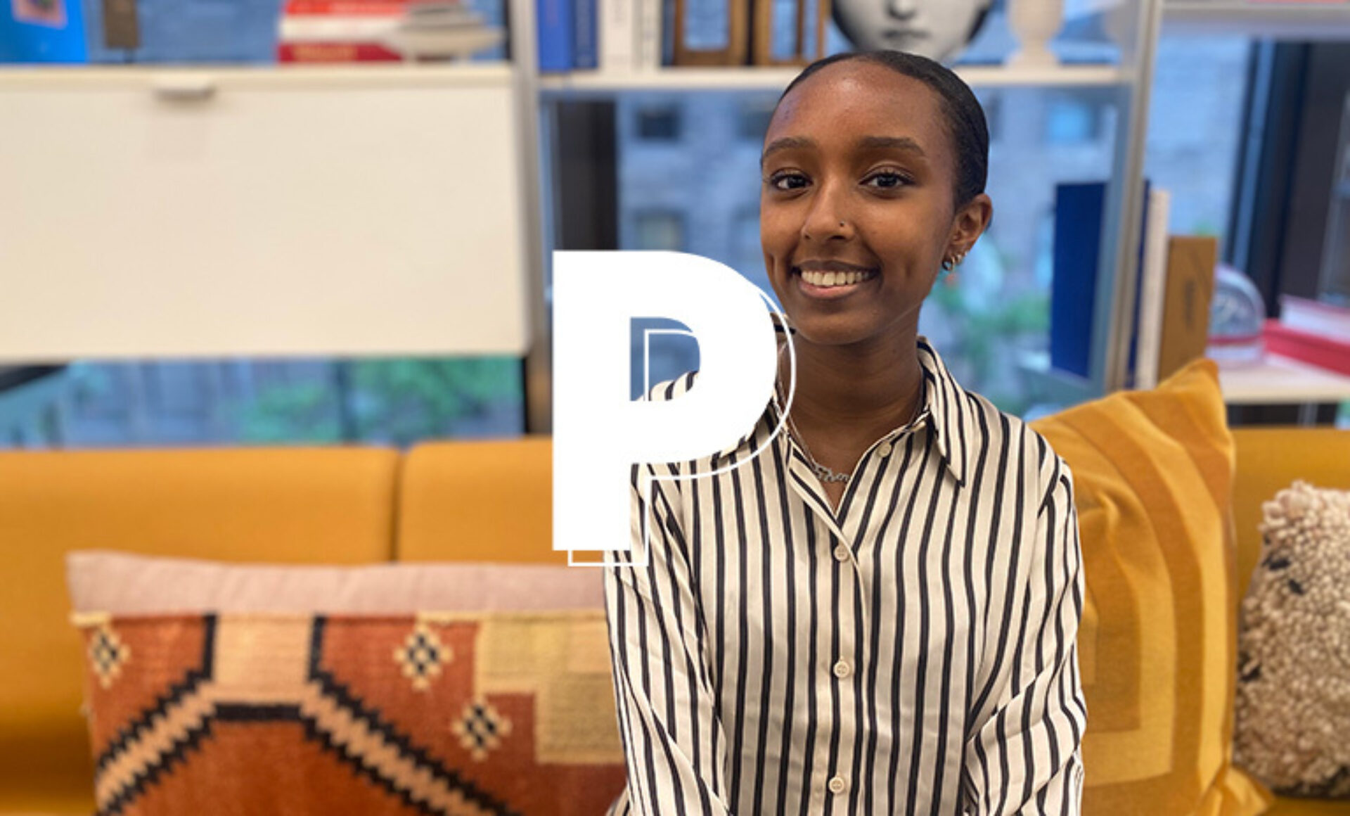 A young woman with dark hair smiles while sitting on a yellow couch, wearing a black and white striped shirt against a bright background.