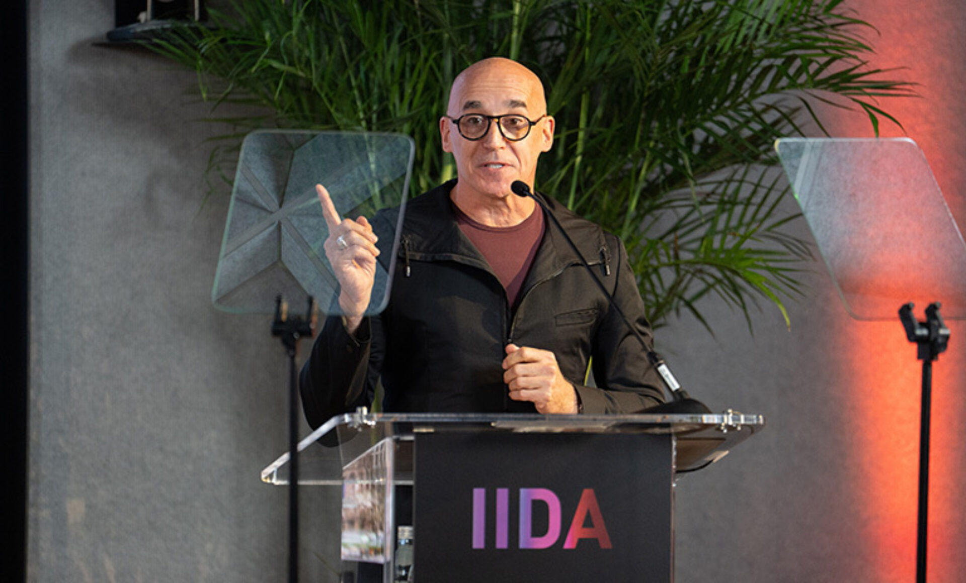 Bald man in glasses and black jacket speaks at a podium labeled IIDA, pointing while teleprompters and a potted palm stand behind him.