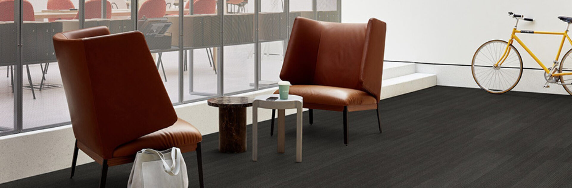 Two brown leather lounge chairs face small wooden and marble side tables holding a takeaway coffee cup, white tote on floor, yellow bike.