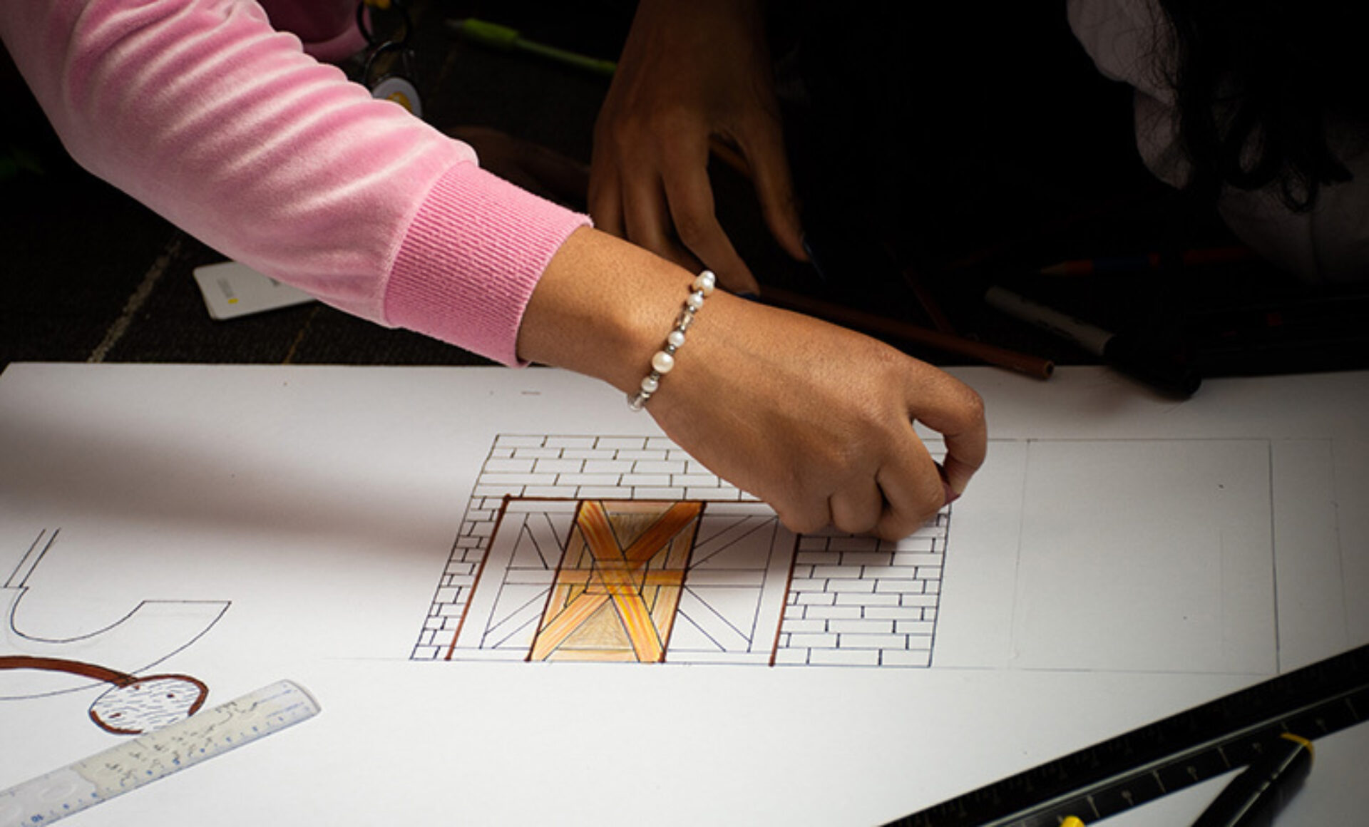 A hand with a bracelet is sketching a detailed design of a door with brick outlines on a large sheet of paper, surrounded by drawing tools.