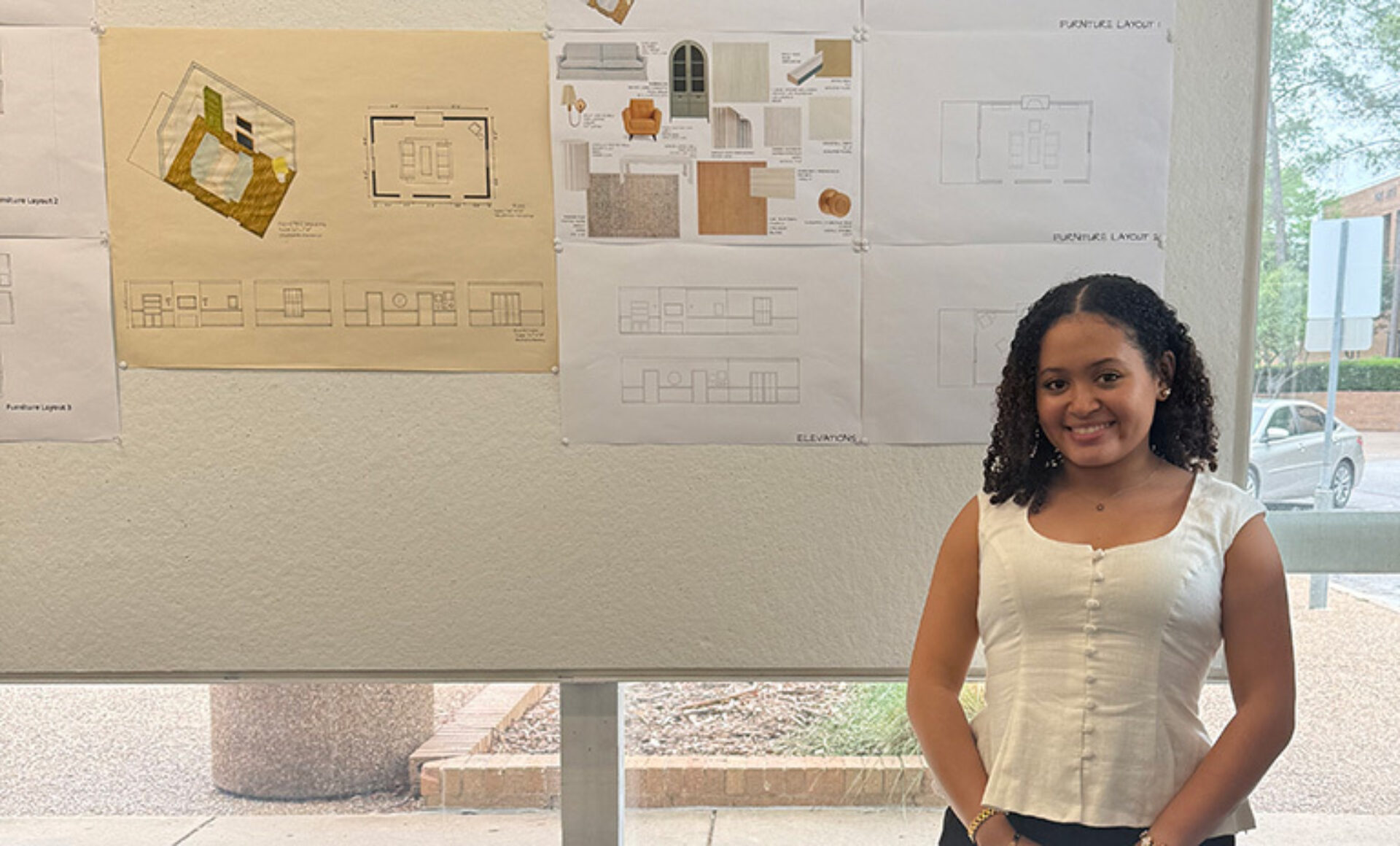 A smiling young woman stands beside pinned architectural drawings on a gallery wall, sunlight from a window revealing brick planter outside.