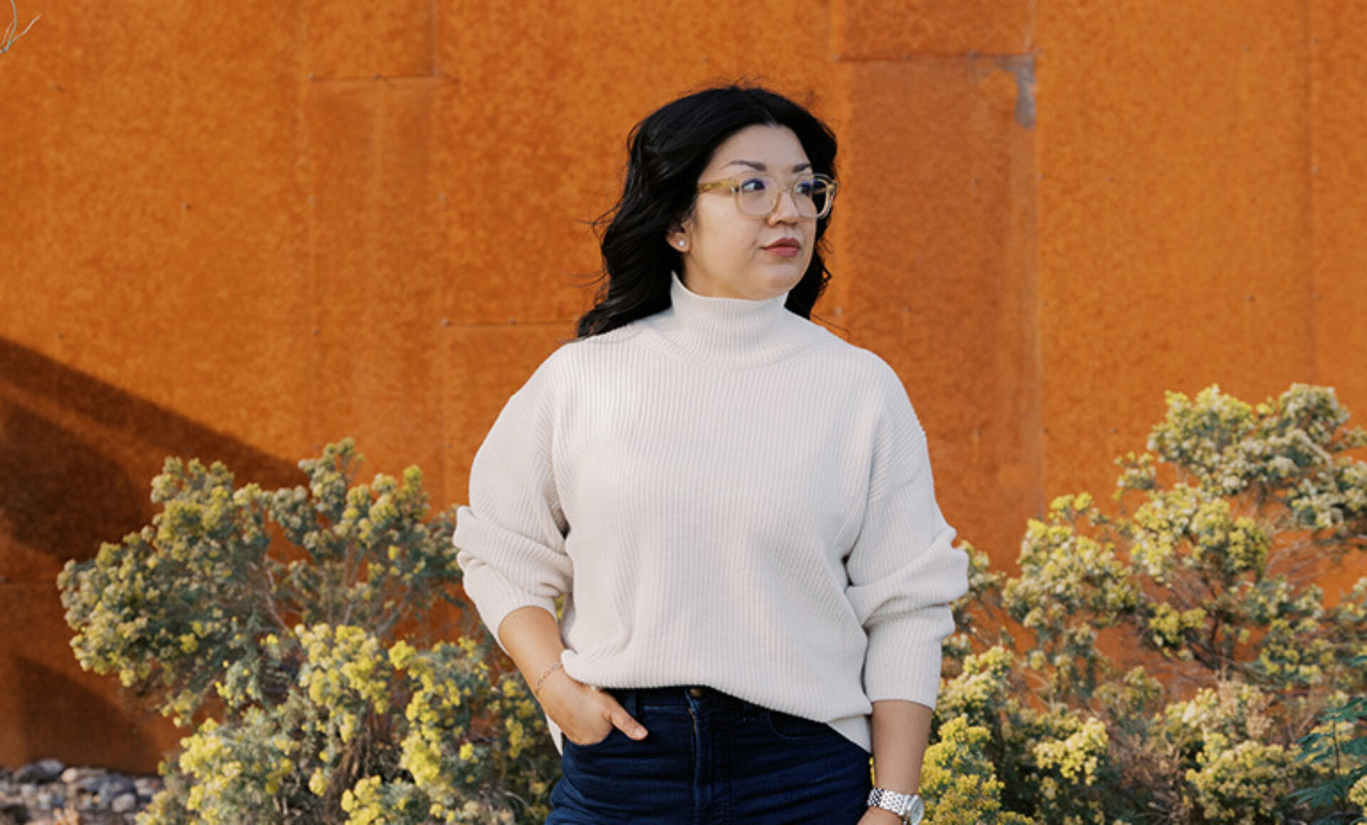 Woman with glasses and black hair in a cream sweater and jeans stands hands in pockets beside yellow shrubs and rust-orange wall.