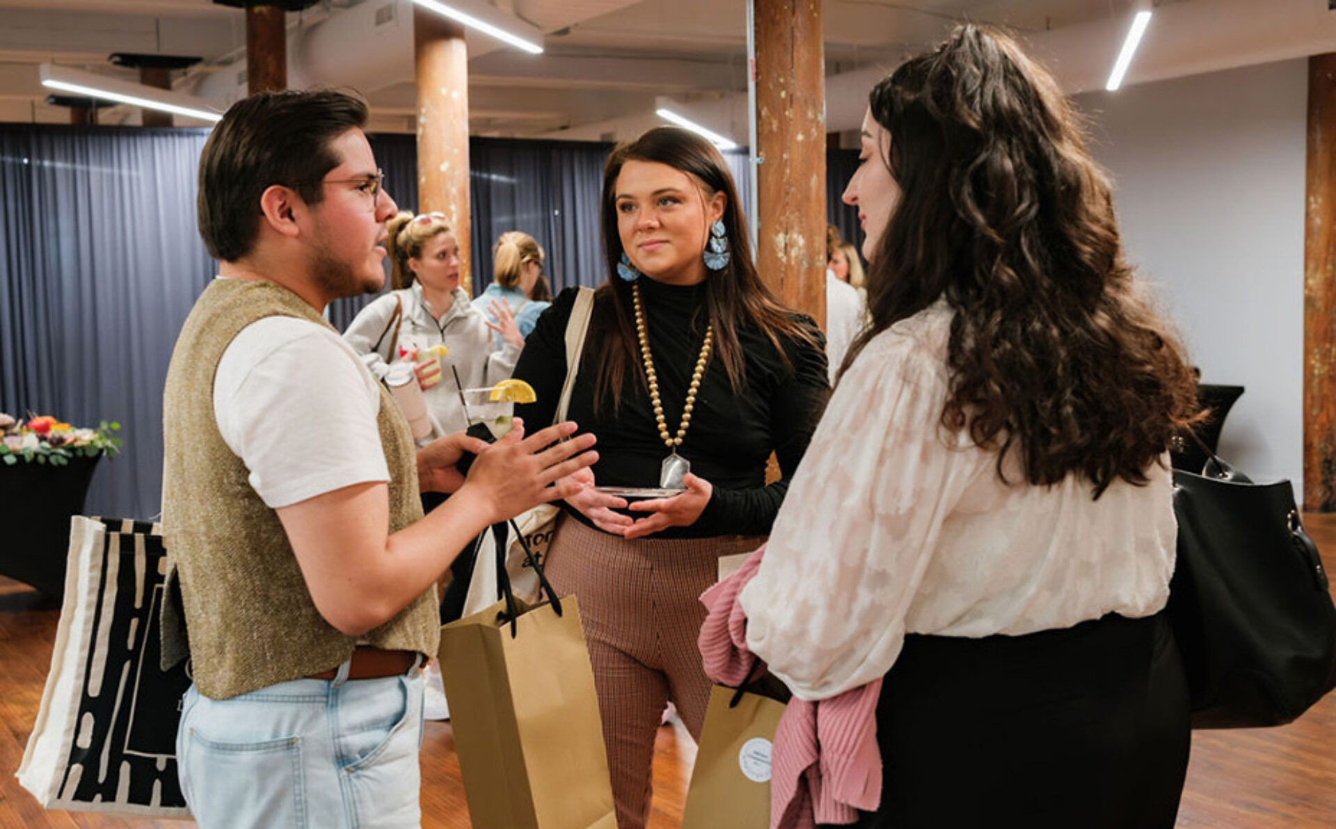 Three individuals engage in conversation at a social event, surrounded by a warm atmosphere and decorative wooden pillars.