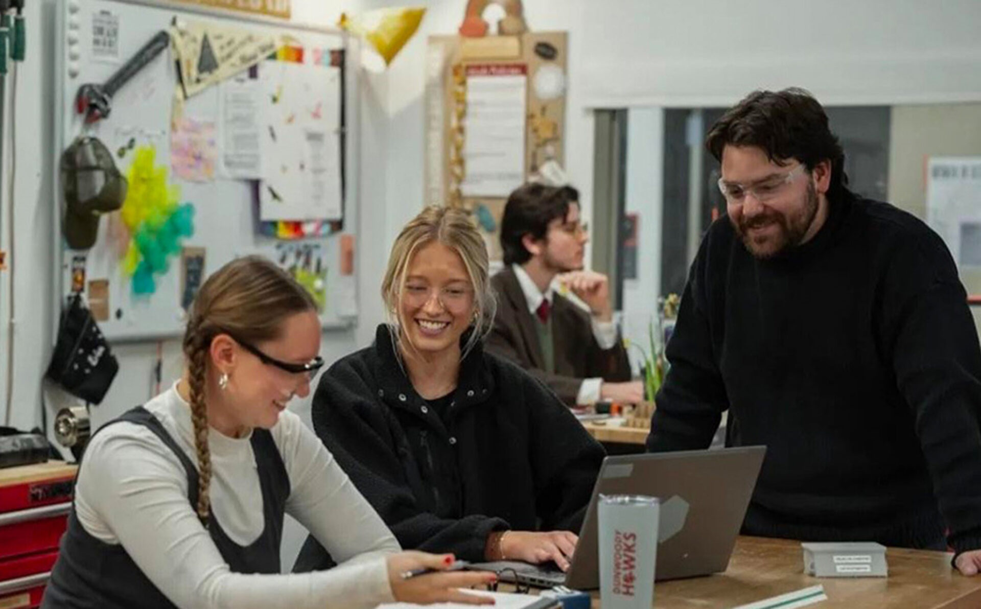 Three people in a workshop collaborate around a laptop: braided woman in safety glasses, a smiling blonde in black and a bearded man leaning in.