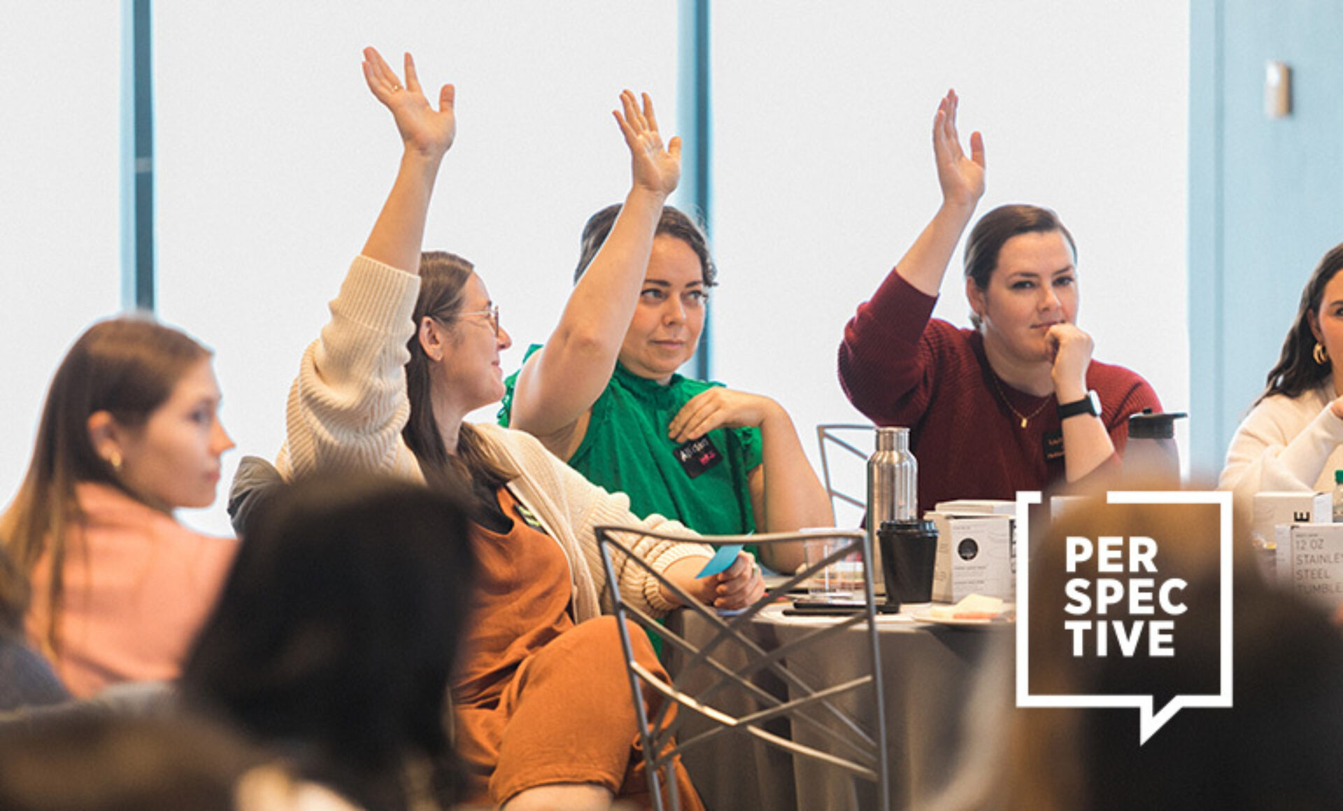 Women seated at a conference round table raise hands to respond, surrounded by notebooks, water bottles, coffee cups and a PERSPECTIVE logo.
