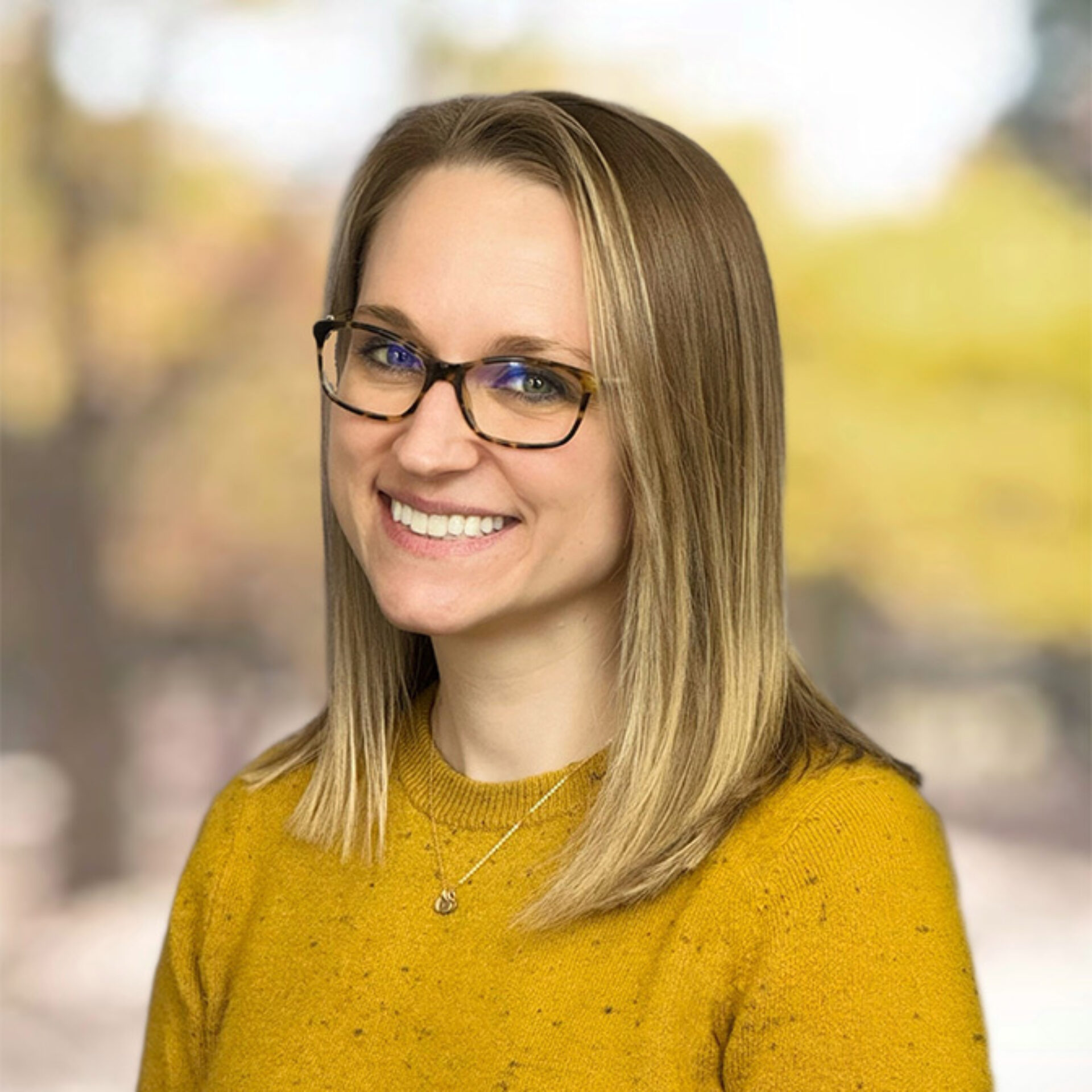 Smiling young woman with straight shoulder-length blonde hair, tortoiseshell glasses and a mustard-yellow sweater, wearing a small pendant.