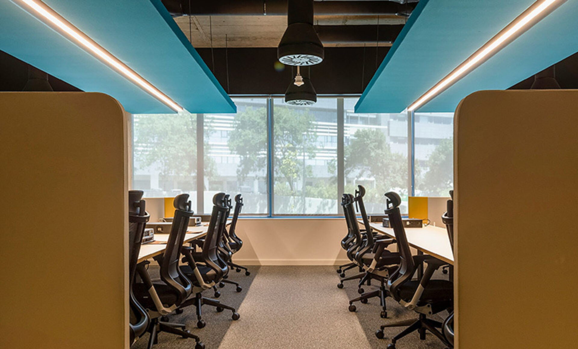 Modern office with two rows of black chairs at long desks beneath teal ceiling panels, pendant lights, and a large window with trees.