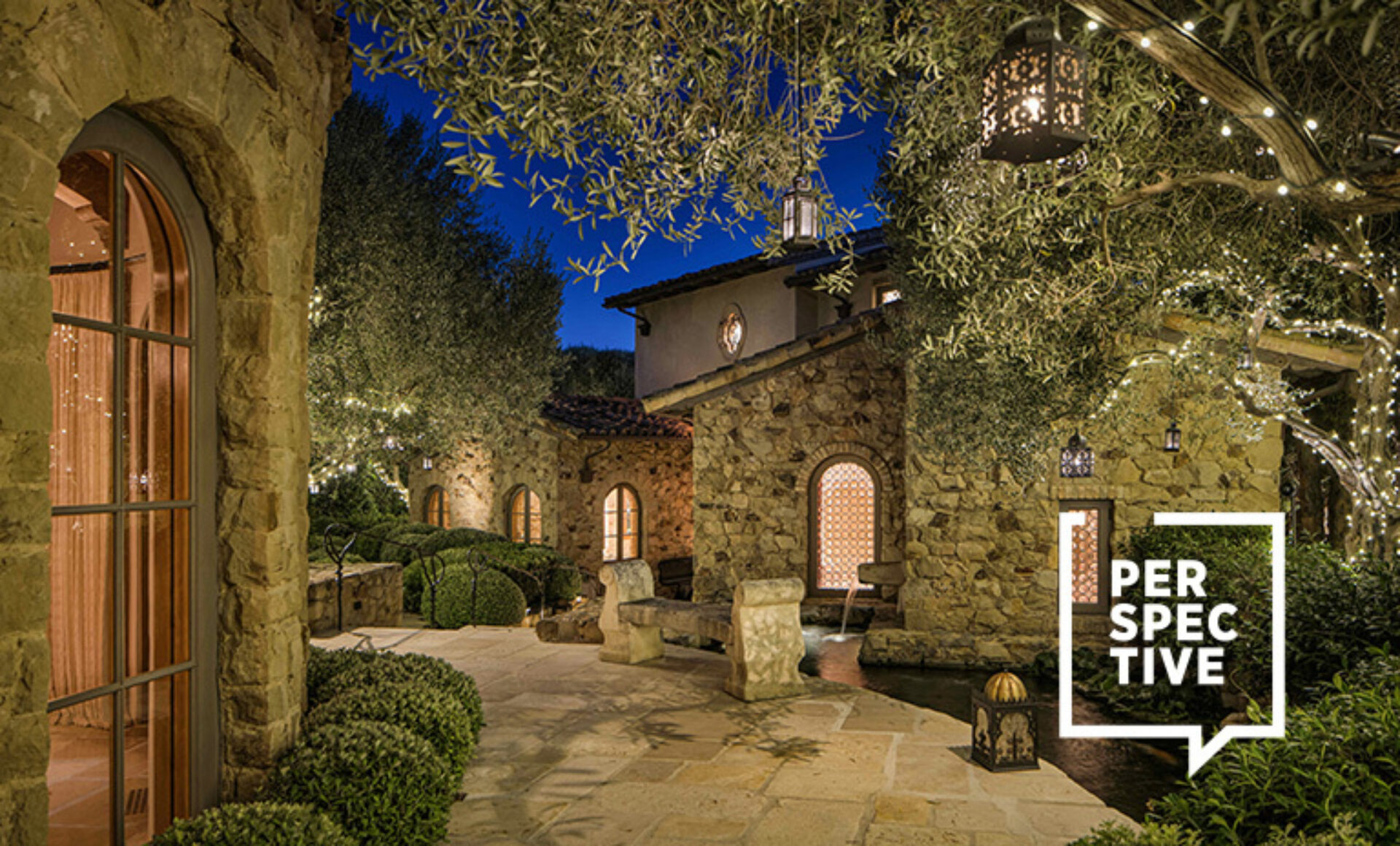 Twilight stone villa with arched windows, olive trees draped in fairy lights, benches by a water channel and a white PERSPECTIVE logo.