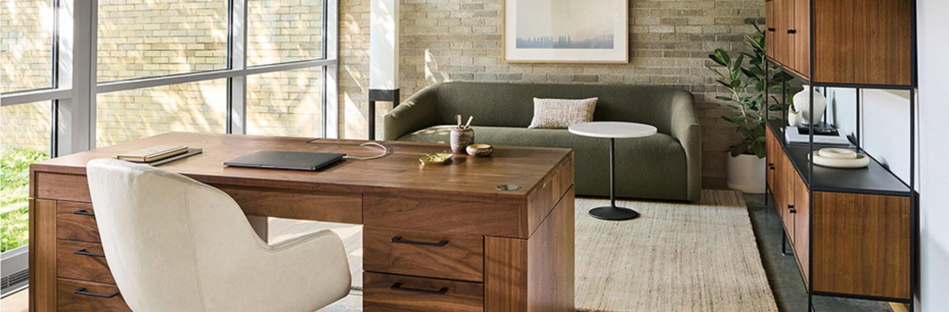 Sunlit modern home office with a walnut desk and cream leather chair facing floor-to-ceiling windows, green sofa, rug, shelving and plants.