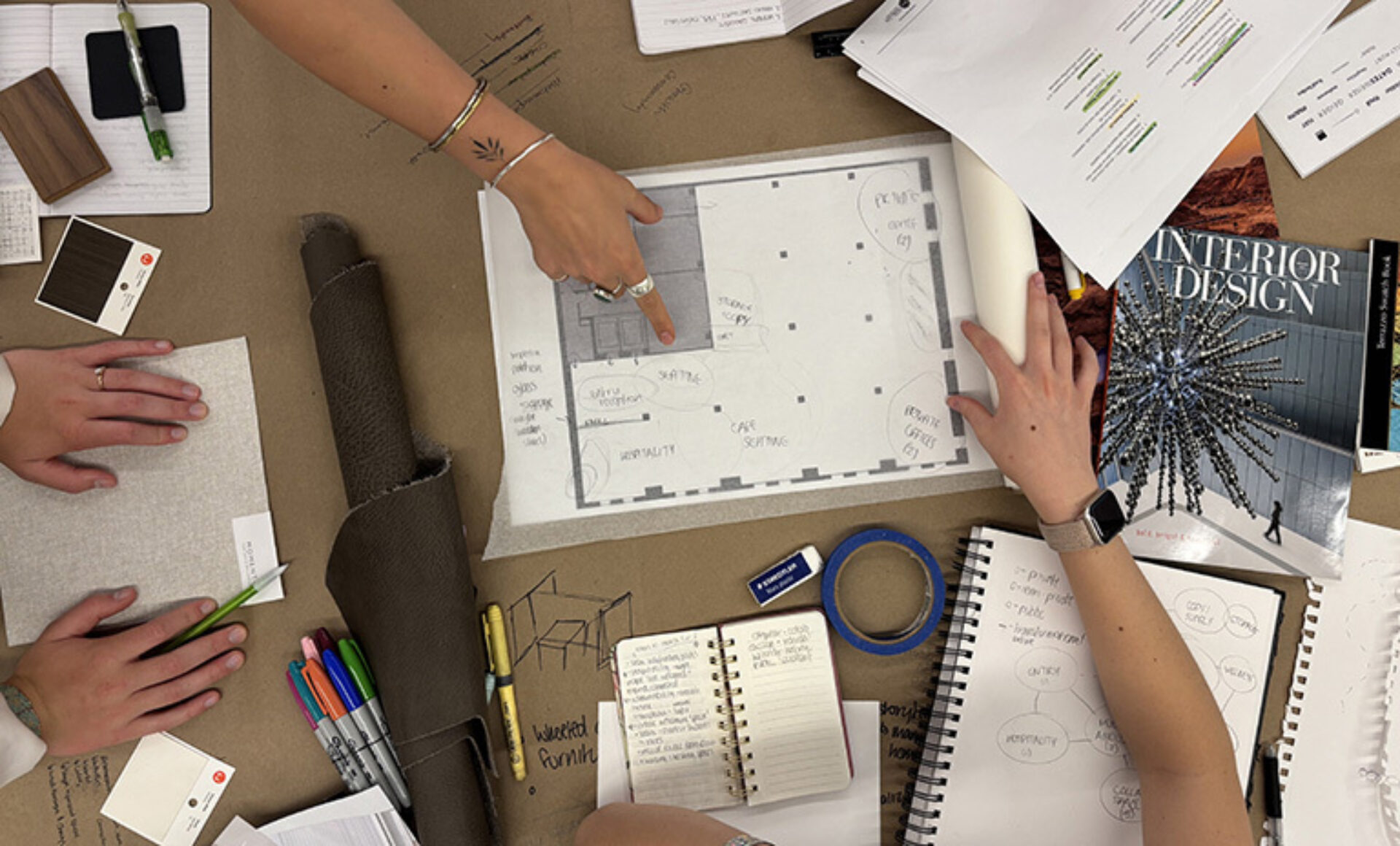Overhead view of designers' hands pointing to a floor plan amid fabric swatches, color chips, markers, notebooks, tape and a design mag.