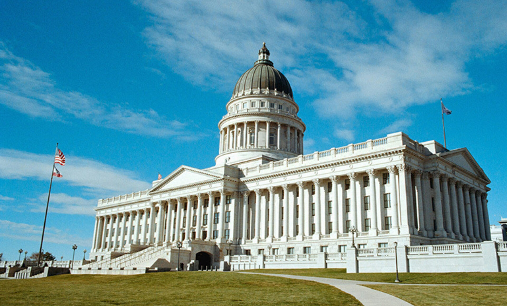 The Utah State Capitol building features a grand dome and classical architecture, surrounded by lush green lawns and a clear sky.
