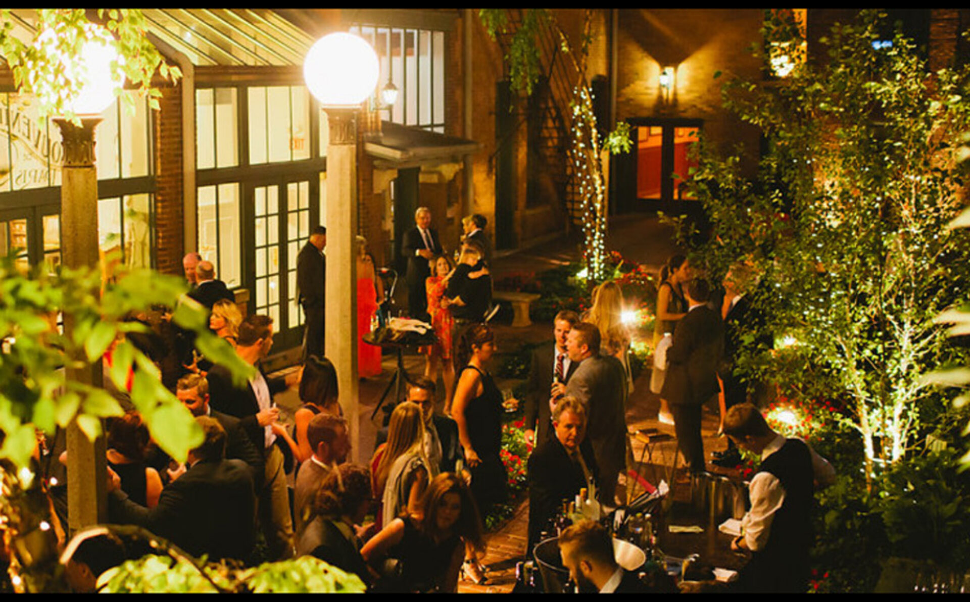 Outdoor evening garden party with well-dressed guests mingling under warm globe lights and fairy-lit trees beside a brick building entrance.