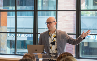 A speaker in a stylish floral shirt gestures enthusiastically while presenting in front of a laptop, engaging an attentive audience.