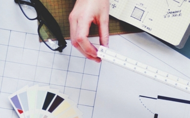 A hand holds a ruler above architectural plans and color swatches, with glasses nearby, indicating design analysis in progress.